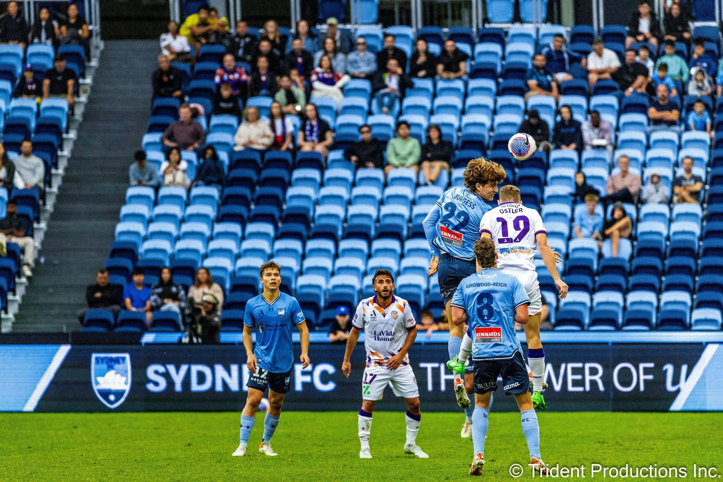 Soccer players competing for a header during a match at Sydney FC stadium, with an audience in the background.