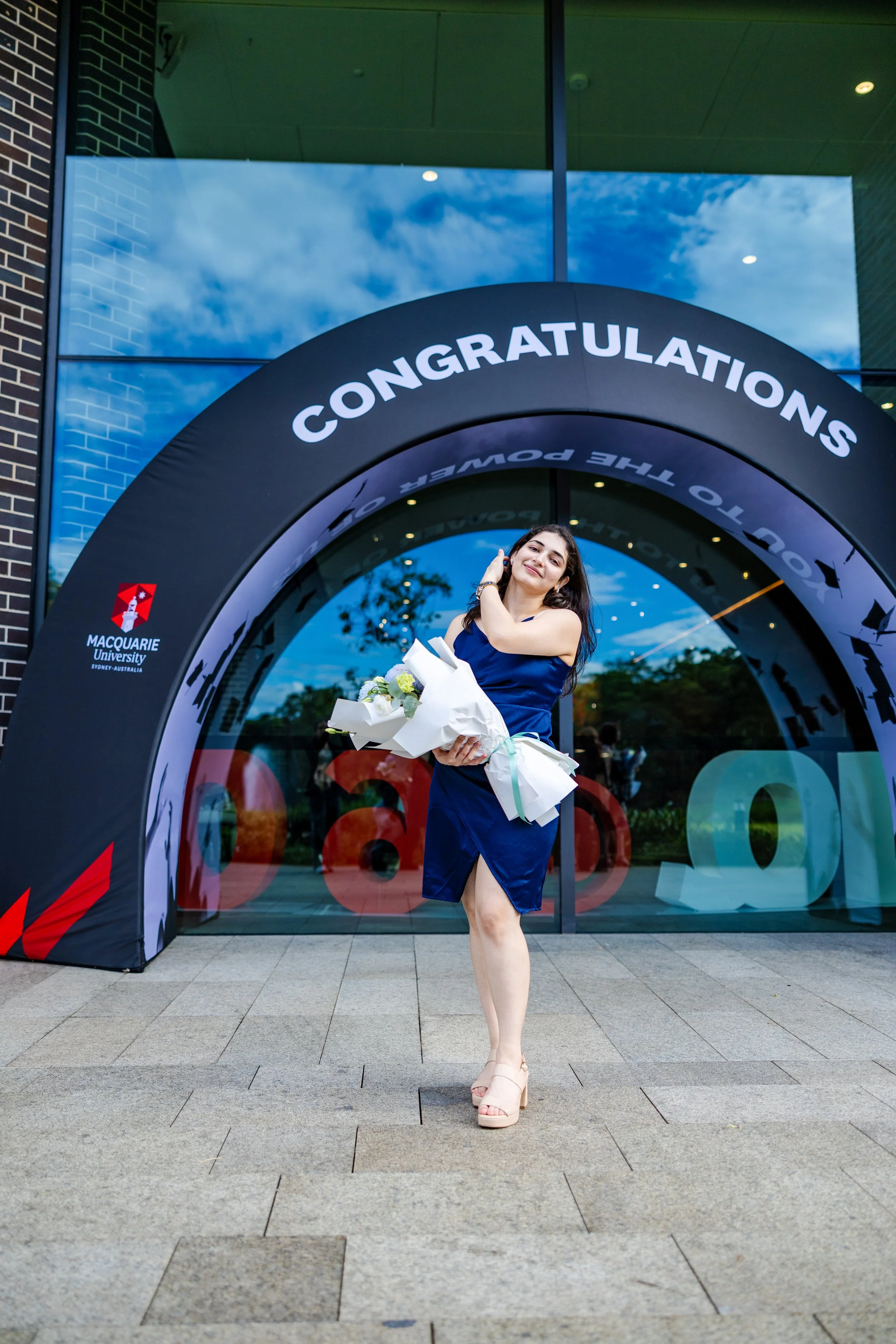 A woman in a blue dress holding a bouquet of flowers stands under a graduation congratulation arch with a school logo in the background, smiling.