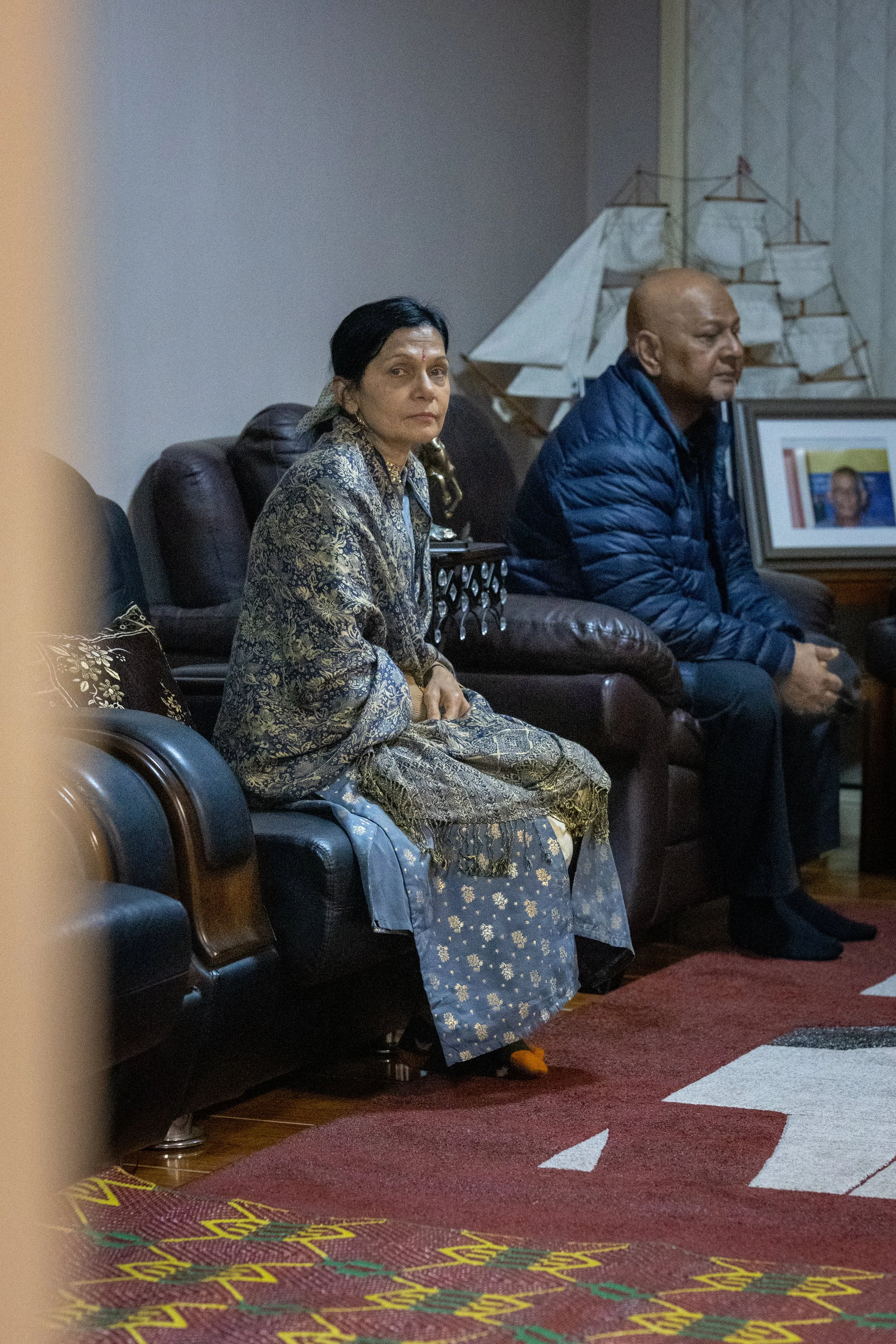 An elderly woman in traditional Indian attire sitting on a black leather sofa, with a bald man in a blue jacket sitting next to her in a living room decorated with model ships and framed photographs.