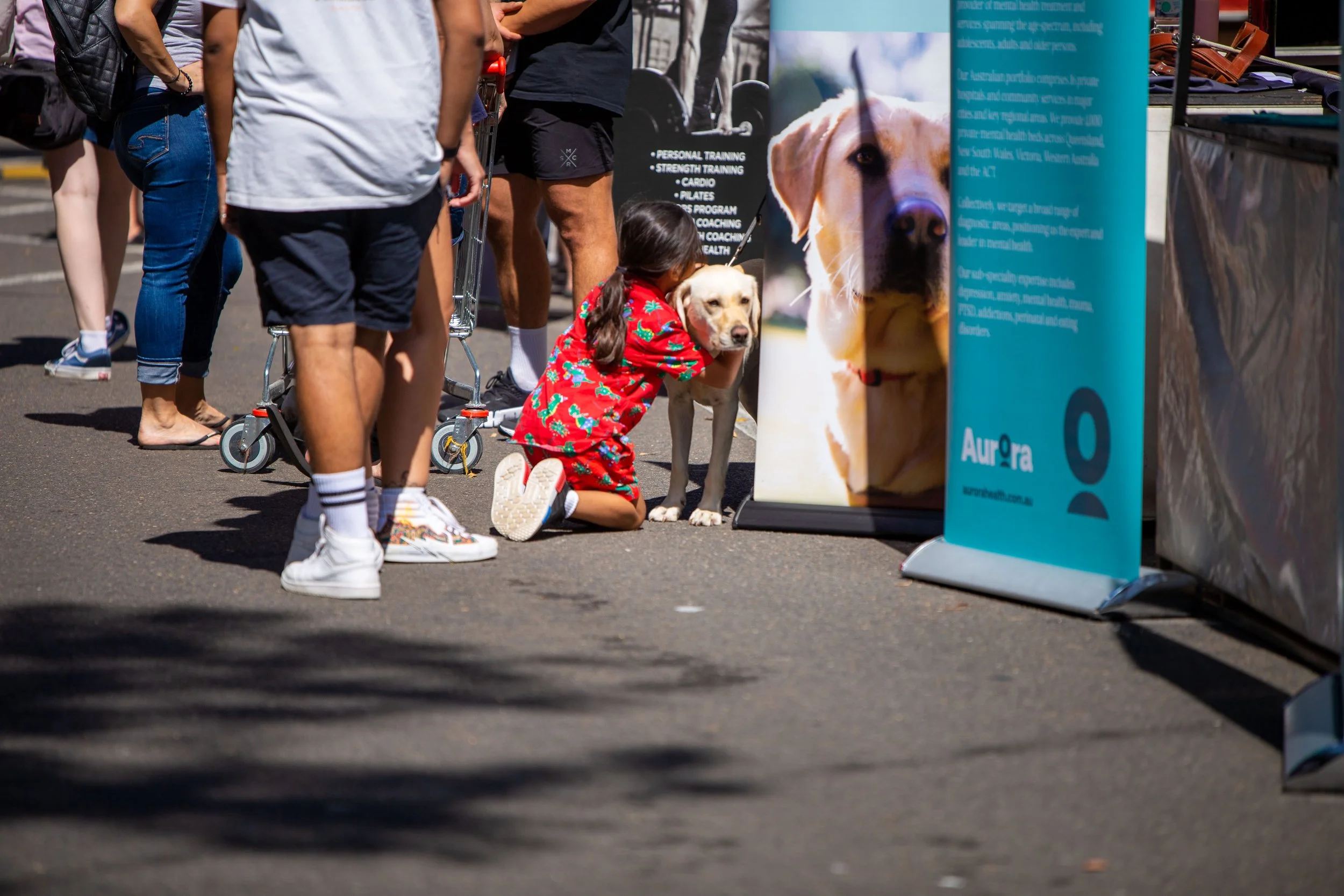 A girl in a red floral dress kneeling and hugging a yellow Labrador retriever dog at an outdoor event or market, with other people standing around and promotional banners nearby.