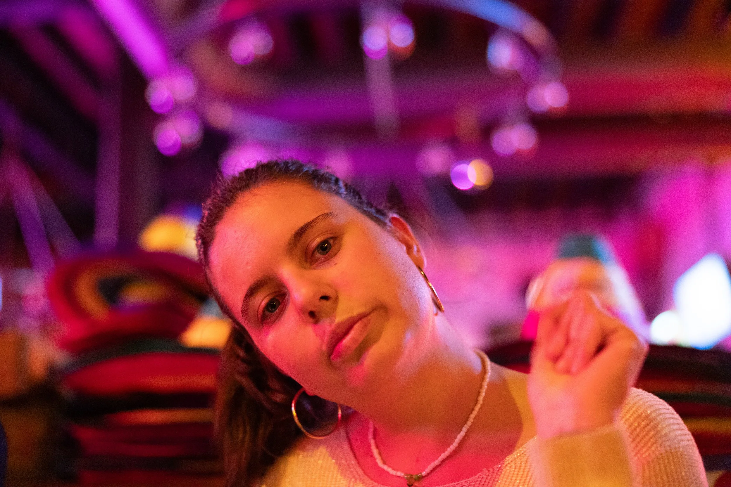 A close-up of a young woman with braided hair, wearing hoop earrings and a pearl necklace, in a colorful, vibrant setting with purple and pink lighting.