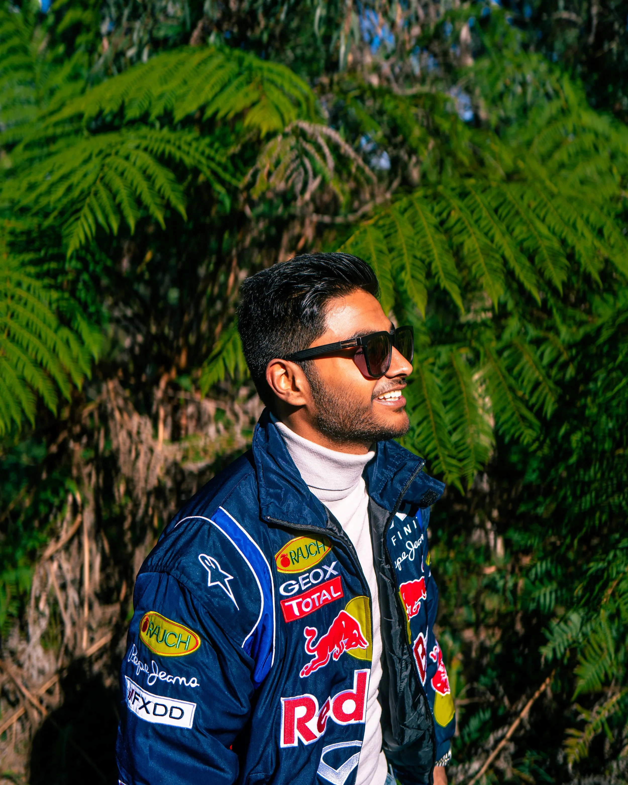 A man wearing sunglasses and a racing jacket with various sponsor patches stands outdoors in front of green fern leaves.