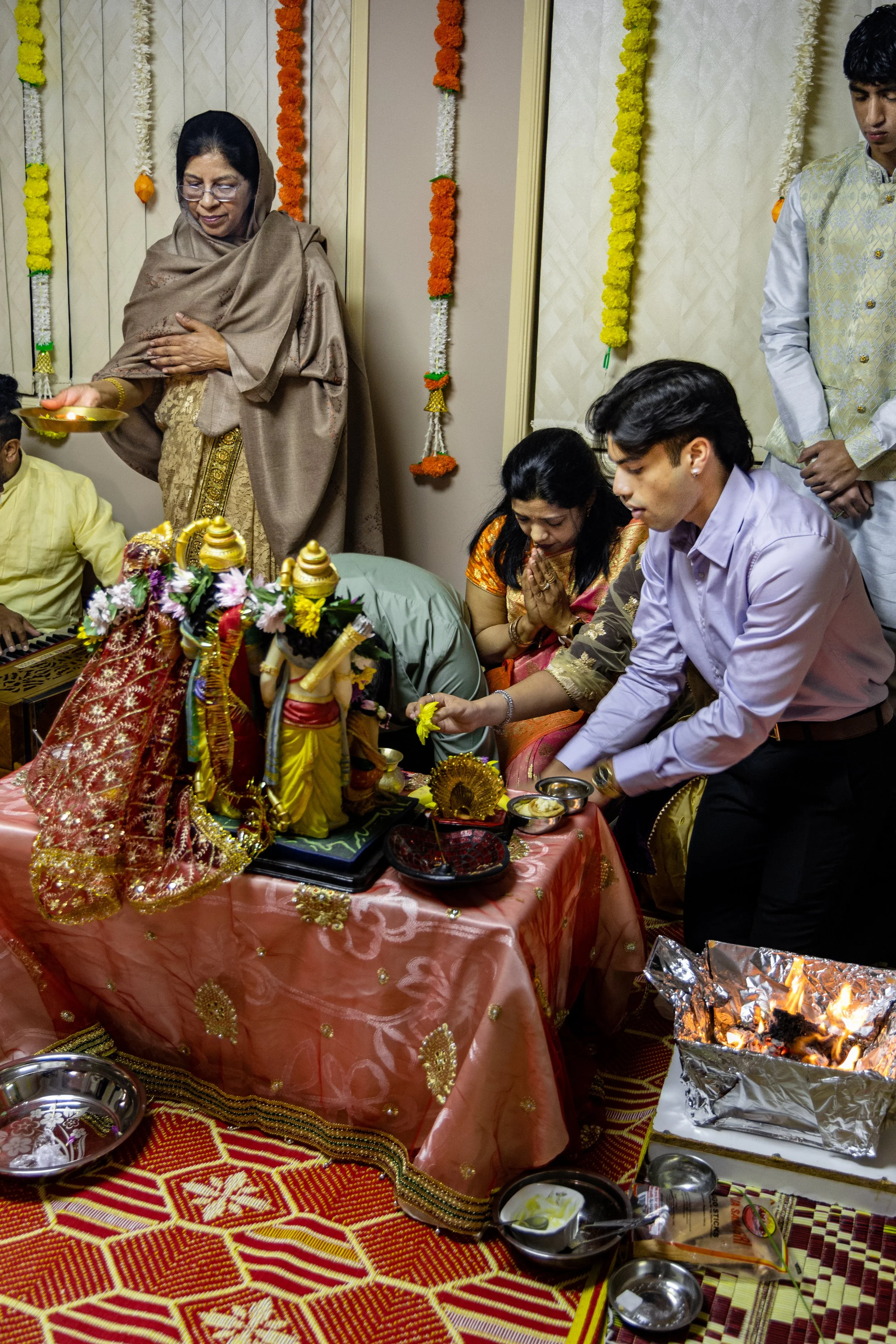People participating in a Hindu religious ceremony with idols and offerings on a table, decorated with flowers and traditional fabrics.