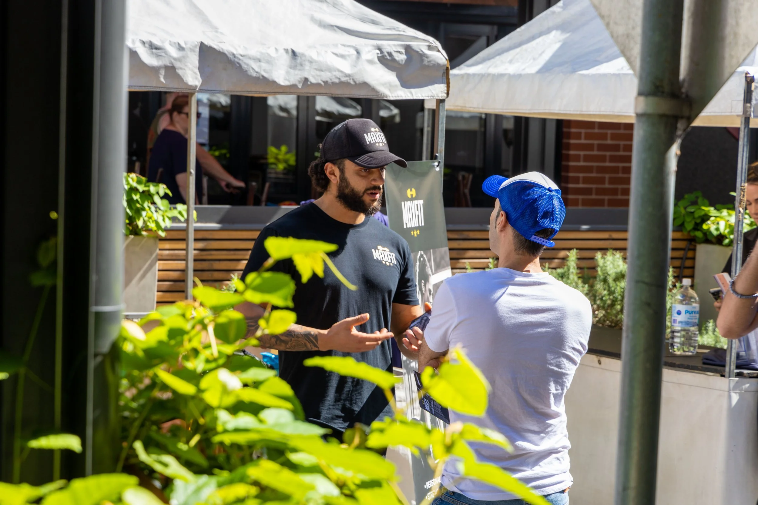 Two men having a conversation outdoors near a food stand. One wears a black cap and T-shirt, the other a blue cap and white T-shirt. There is greenery in the foreground and a water bottle on the stand.