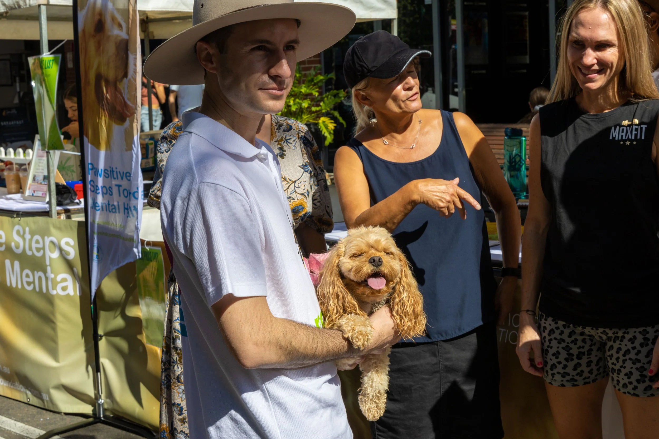 A man wearing a white polo shirt and large floppy hat holding a small, curly-haired dog with its tongue out. Two women stand nearby, one in a blue sleeveless top and black cap pointing, the other in a black sleeveless shirt and leopard print shorts, 
