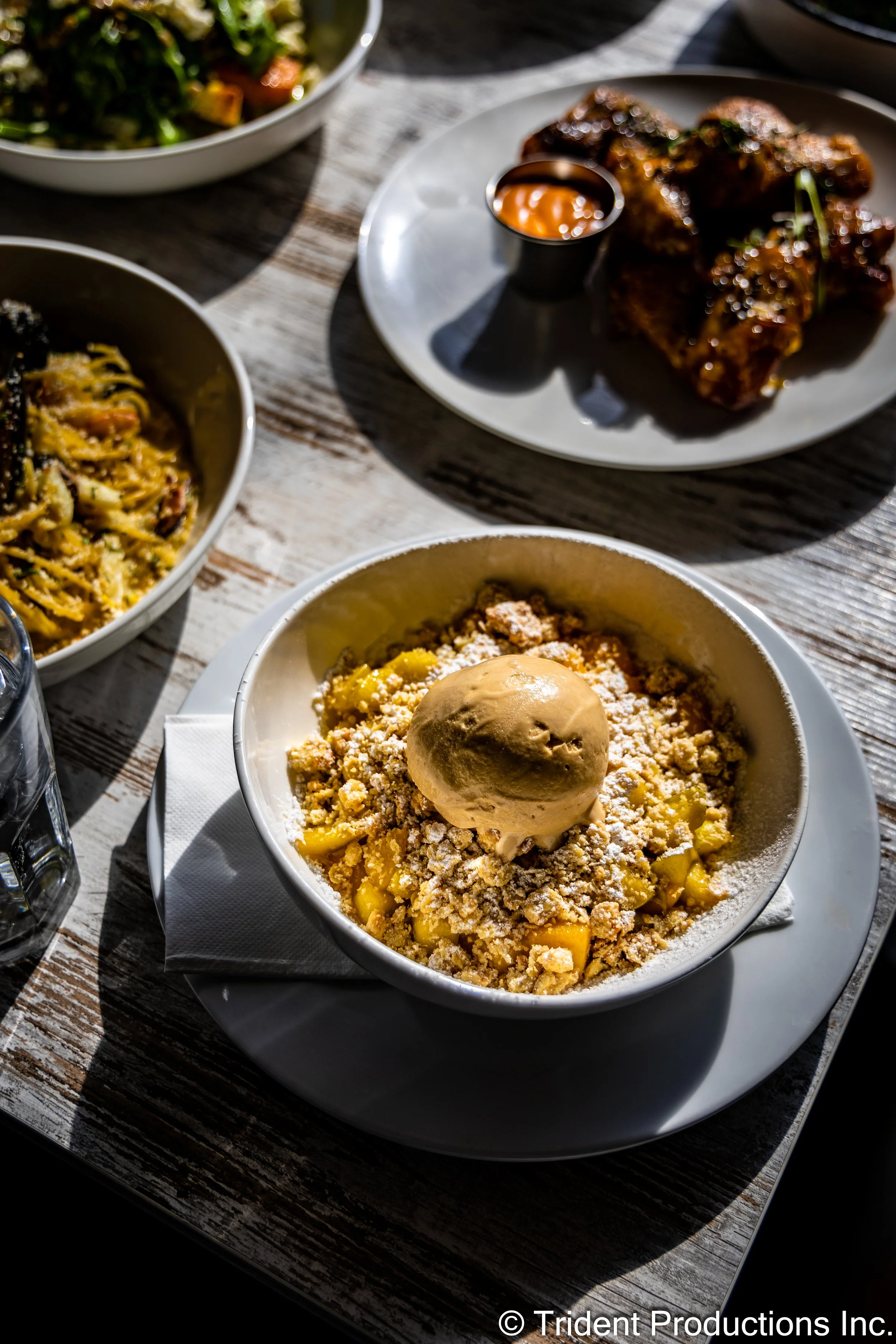 A bowl of apple crumble topped with a scoop of ice cream, with a meal of ribs and pasta dishes on a rustic table.