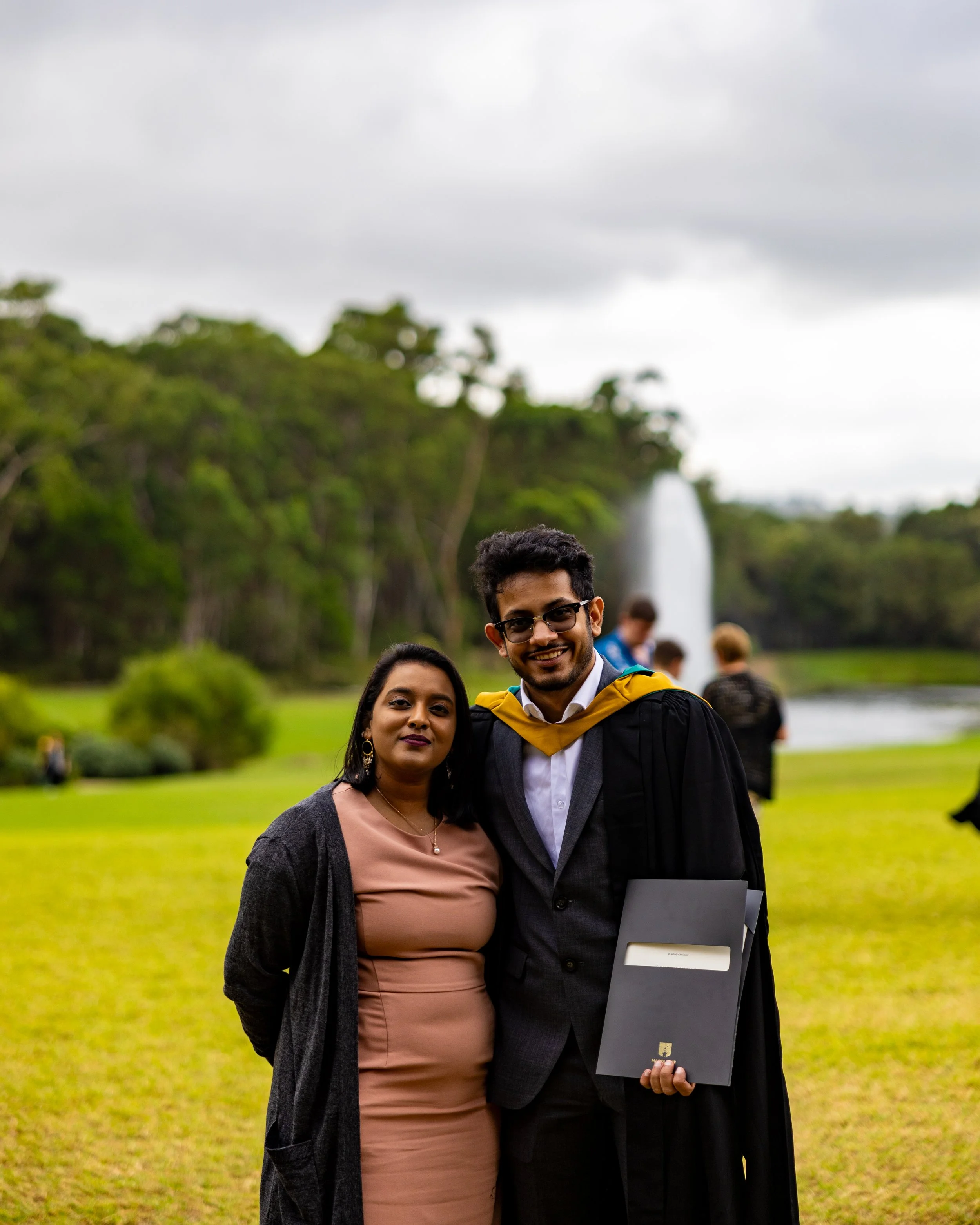A smiling man in graduation attire standing next to a woman outdoors in front of a fountain and trees.