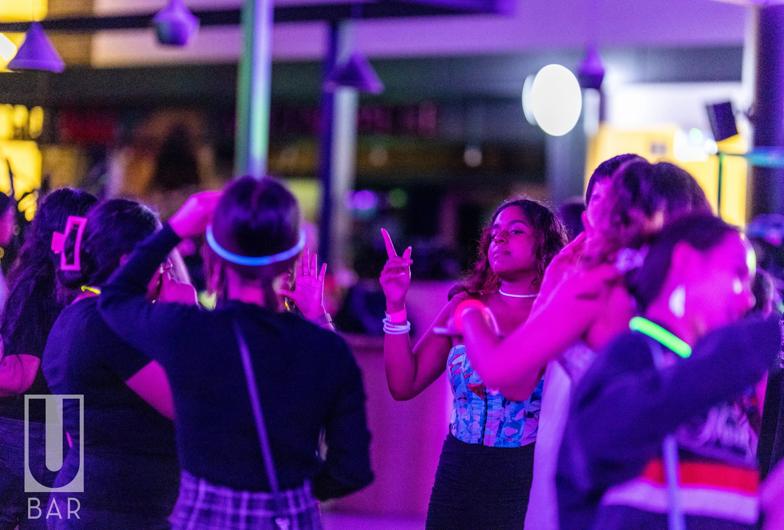 Group of young people dancing and socializing at a nightclub with neon lighting, wearing glow accessories, and enjoying the night.