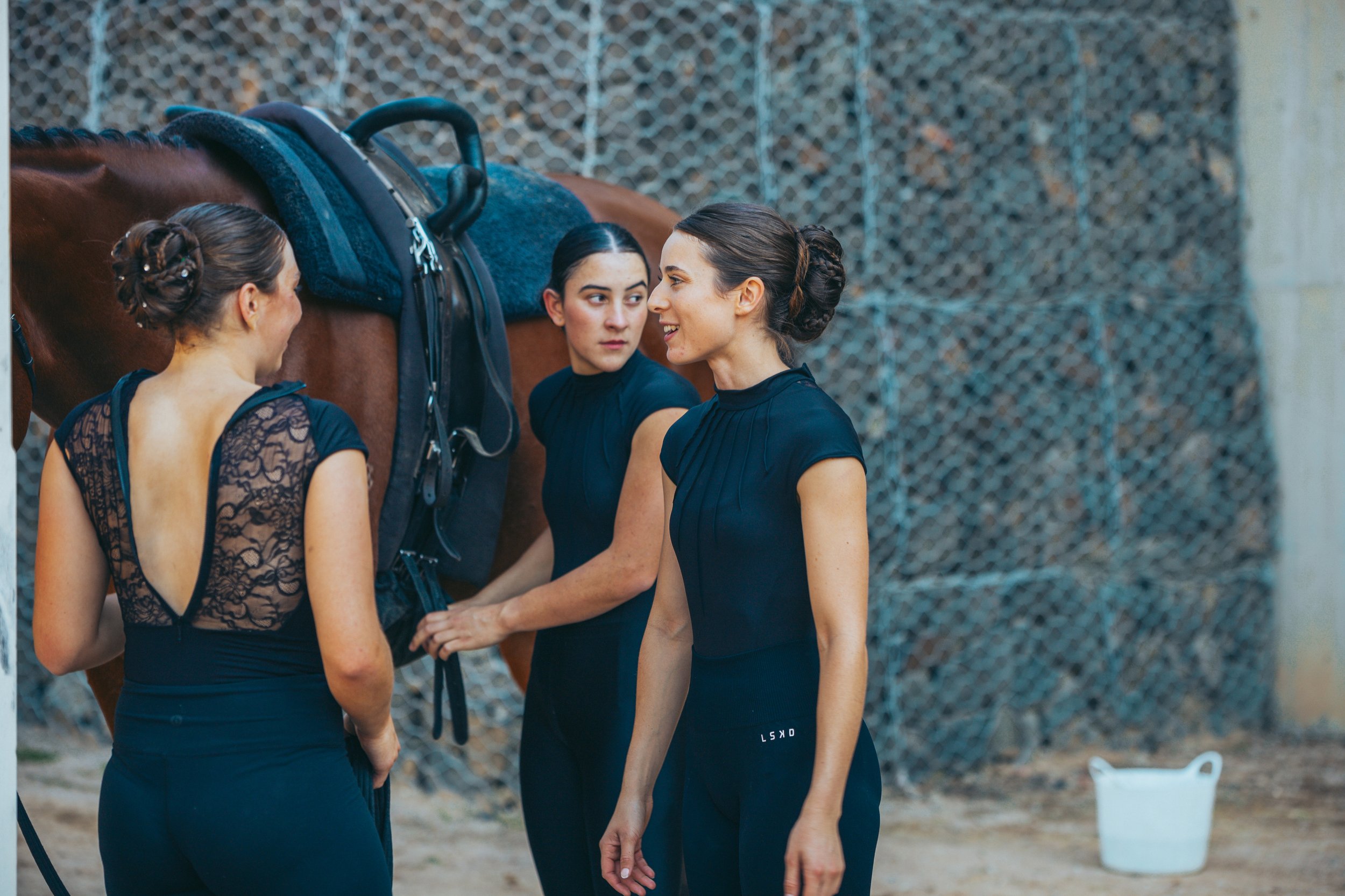 Three women in black outfits speaking near a brown horse with a saddle, against a chain-link fence background.