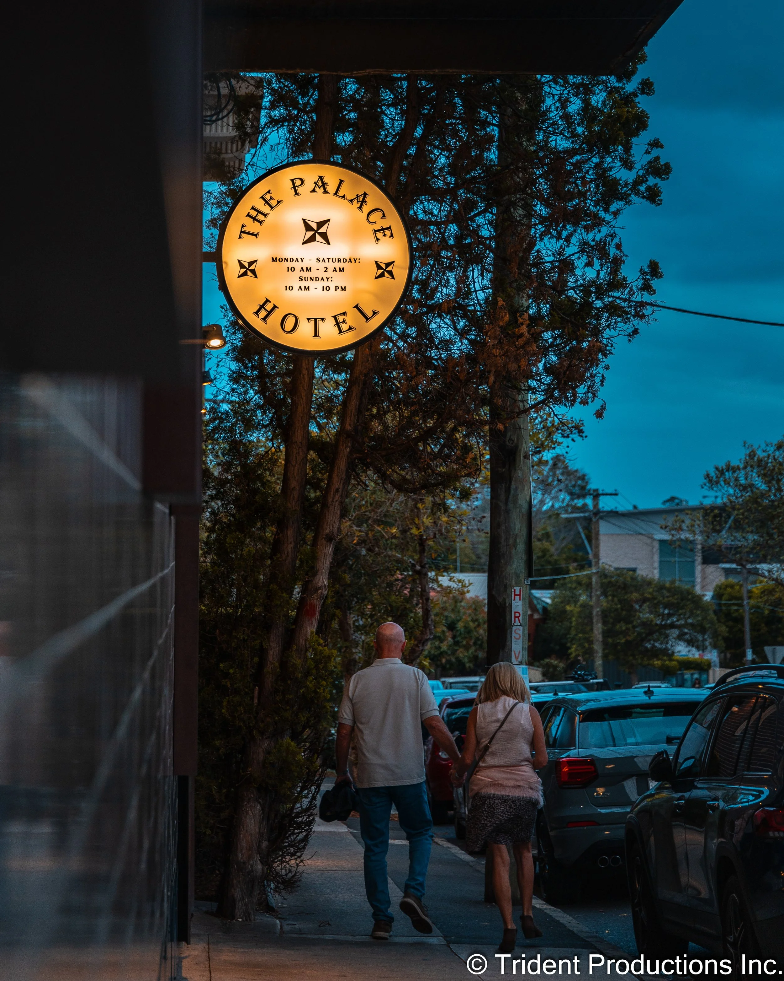 Two people walk hand in hand along a sidewalk near parked cars and a lit hotel sign that reads 'The Palace Hotel' with hours of operation listed.