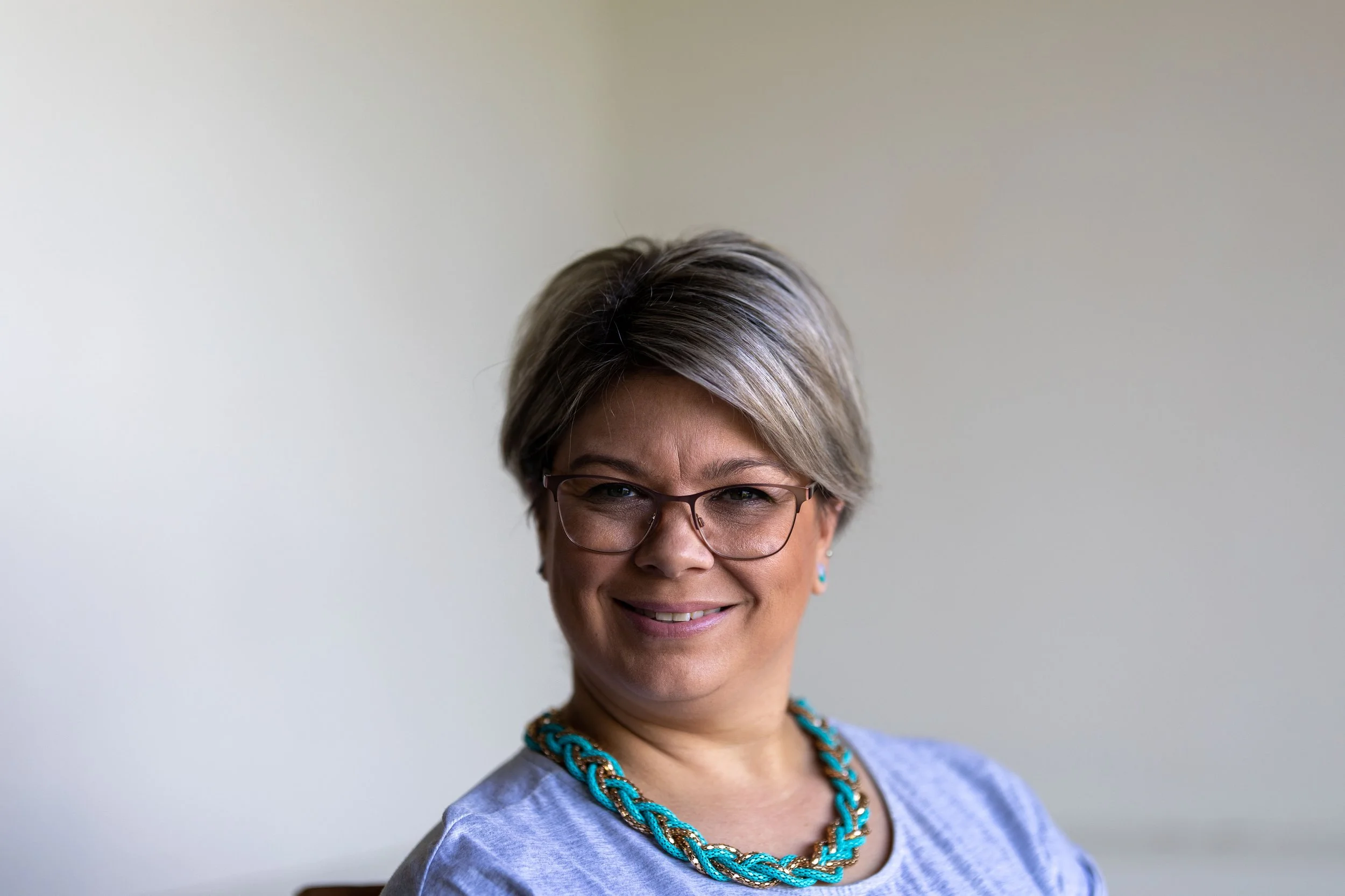 A middle-aged woman with short gray hair, wearing glasses, a turquoise and gold necklace, and a light gray shirt, smiling at the camera against a plain light background.