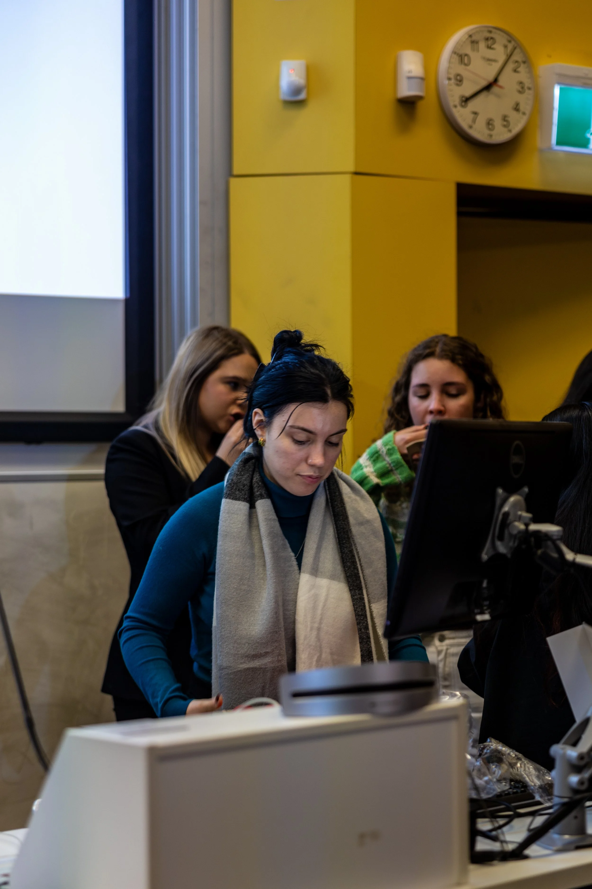 Group of young women working at a computer in a classroom or lecture hall, with a yellow wall, clock, and projection screen in the background.
