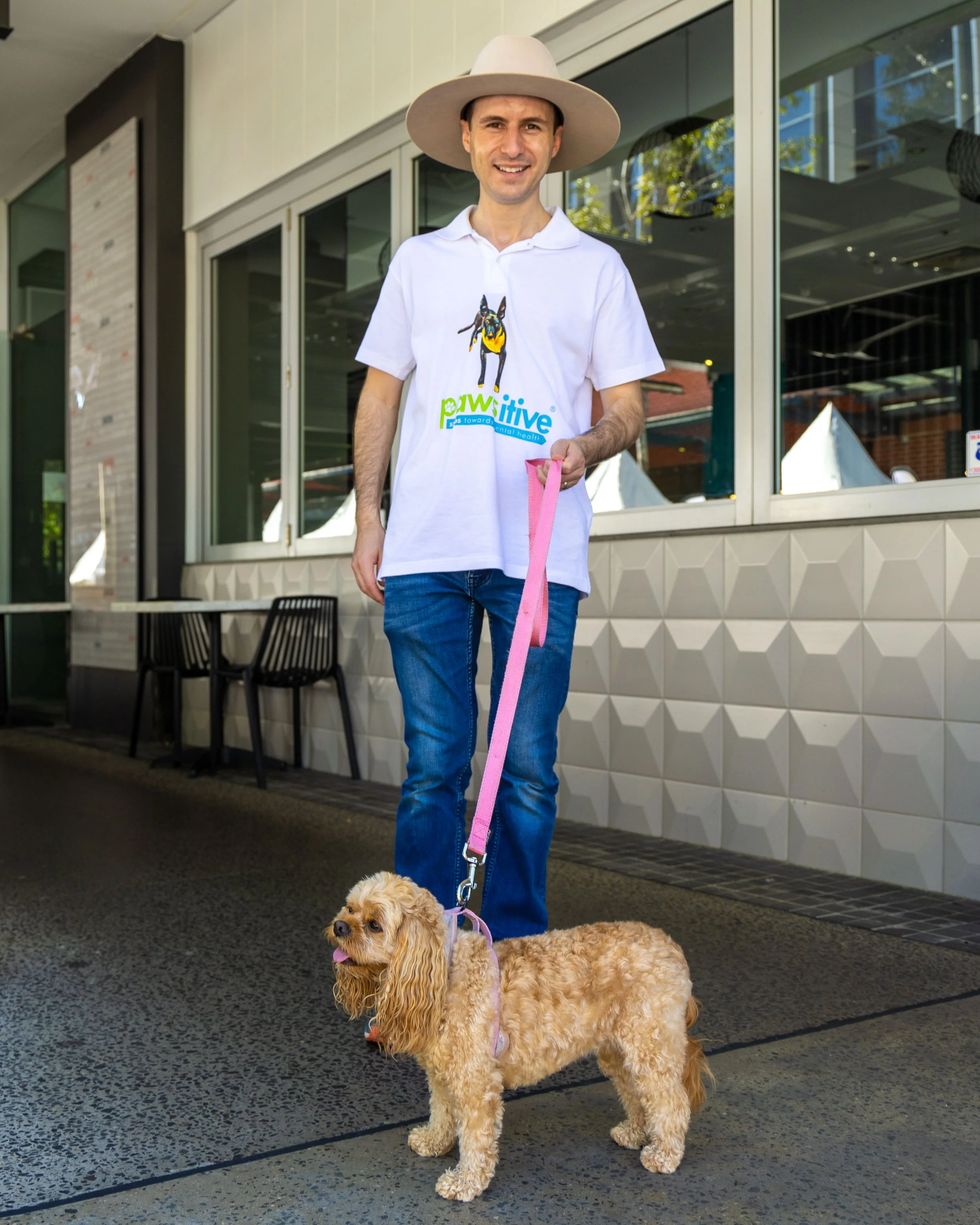 A man smiling and holding a pink leash attached to a small tan-colored dog with long ears, standing outdoors in front of a building with large windows.