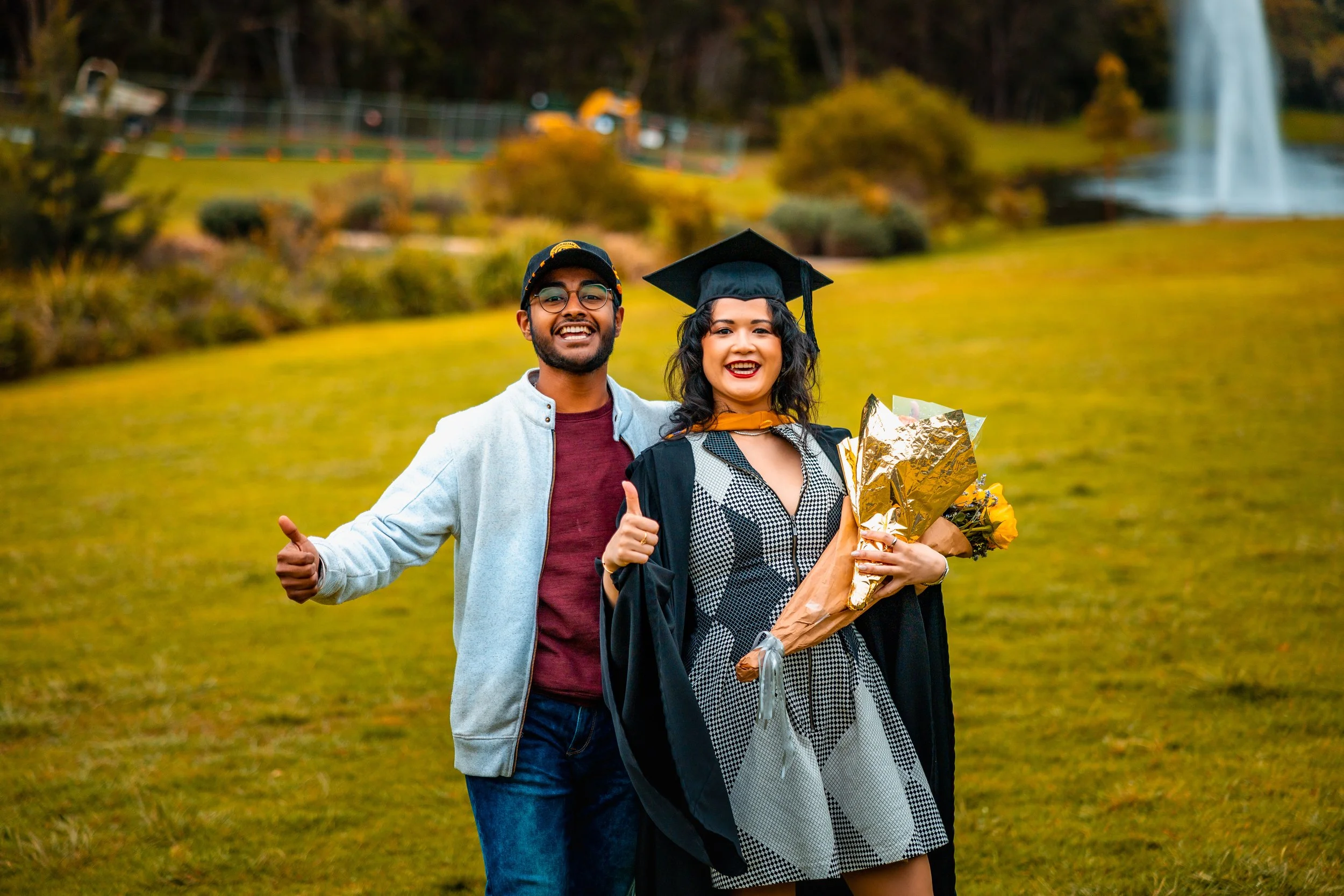 A graduate woman in a black gown and cap holding a bouquet of flowers, smiling, with a happy man giving a thumbs up in a park with a fountain in the background.