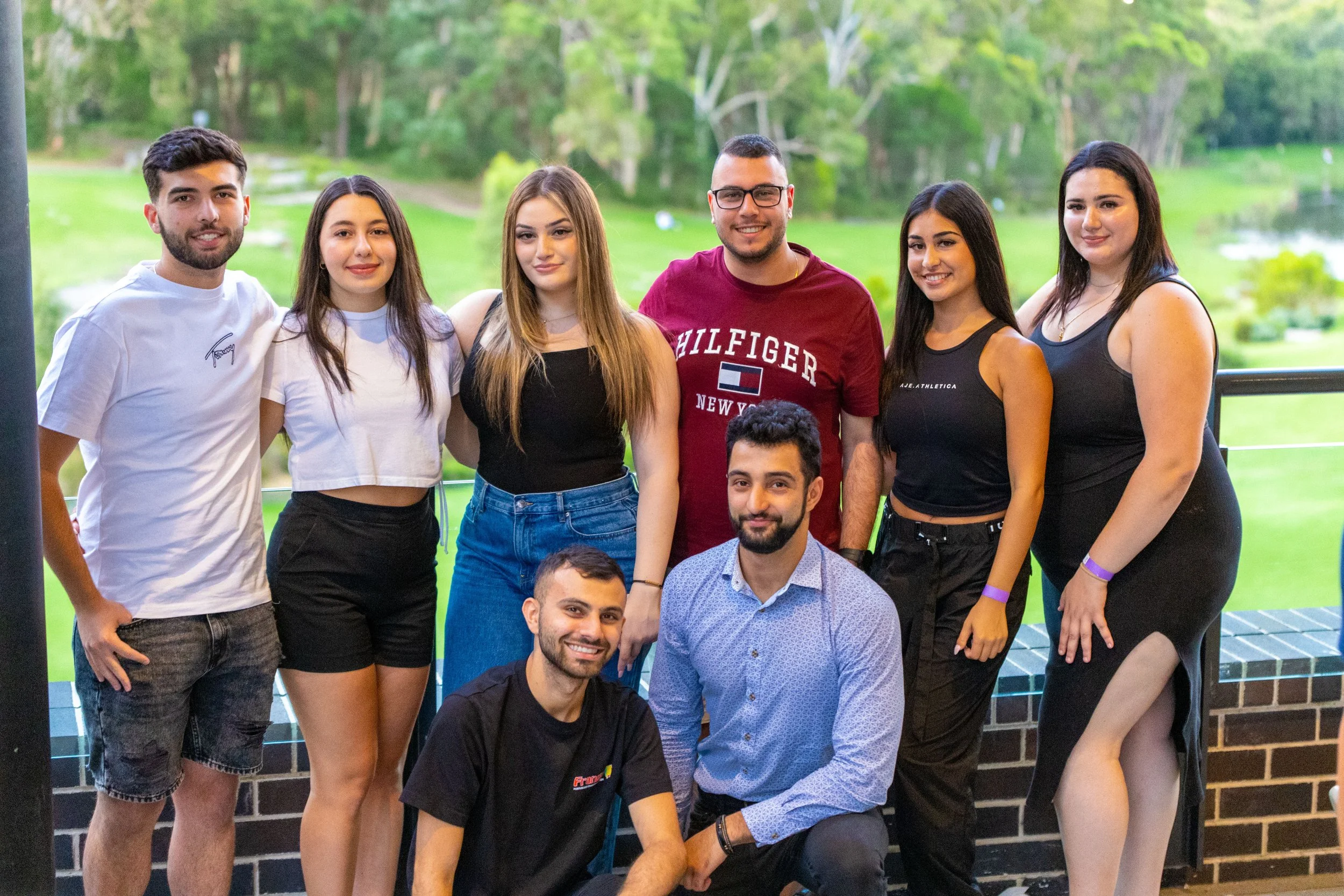 Group of nine young adults posing outdoors with greenery and water in the background.