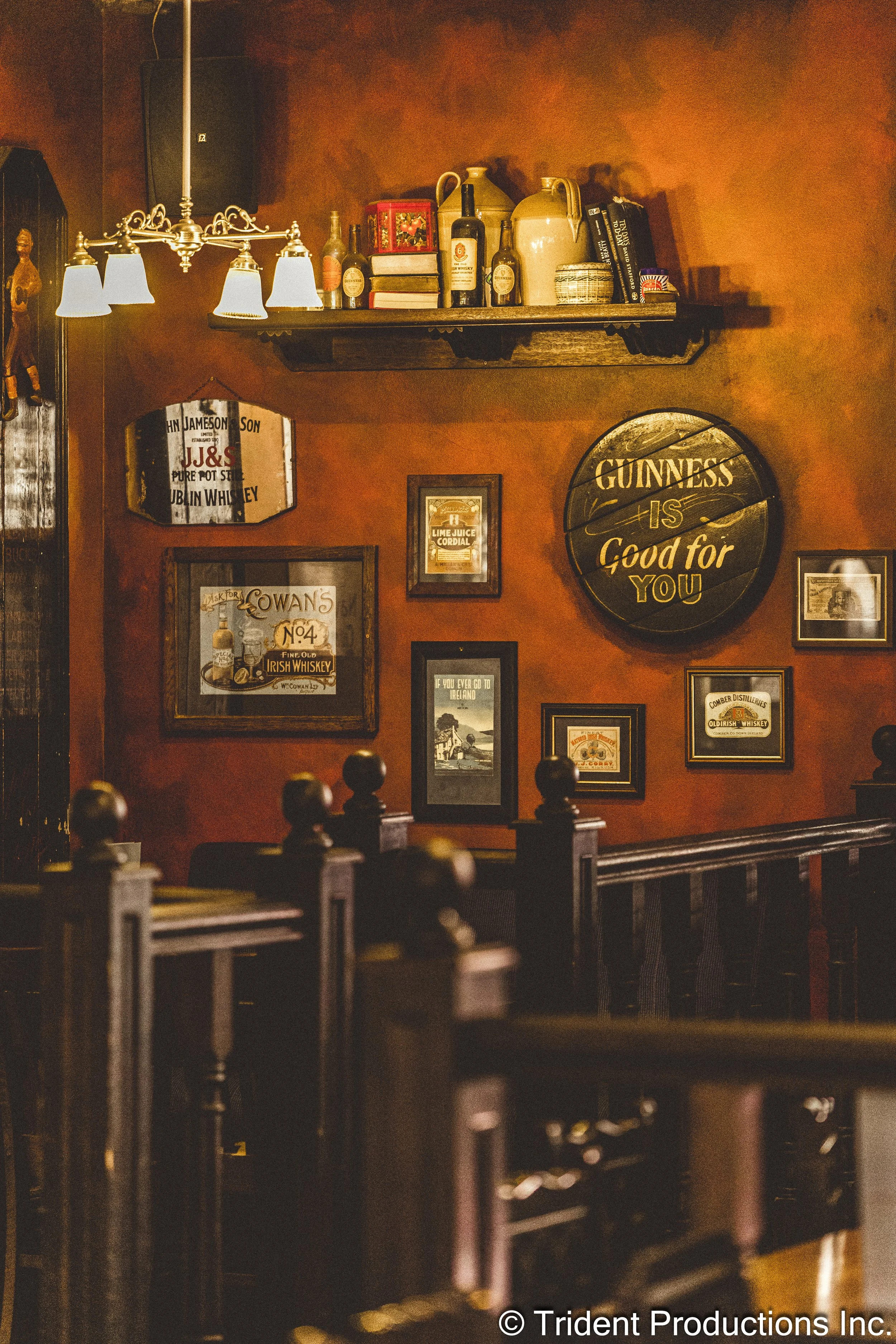 Interior of a cozy pub with wall decorations including framed posters and a large round sign reading 'GUINNESS IS Good for You'. Wooden chairs and a shelf with bottles and decorative items are also visible.