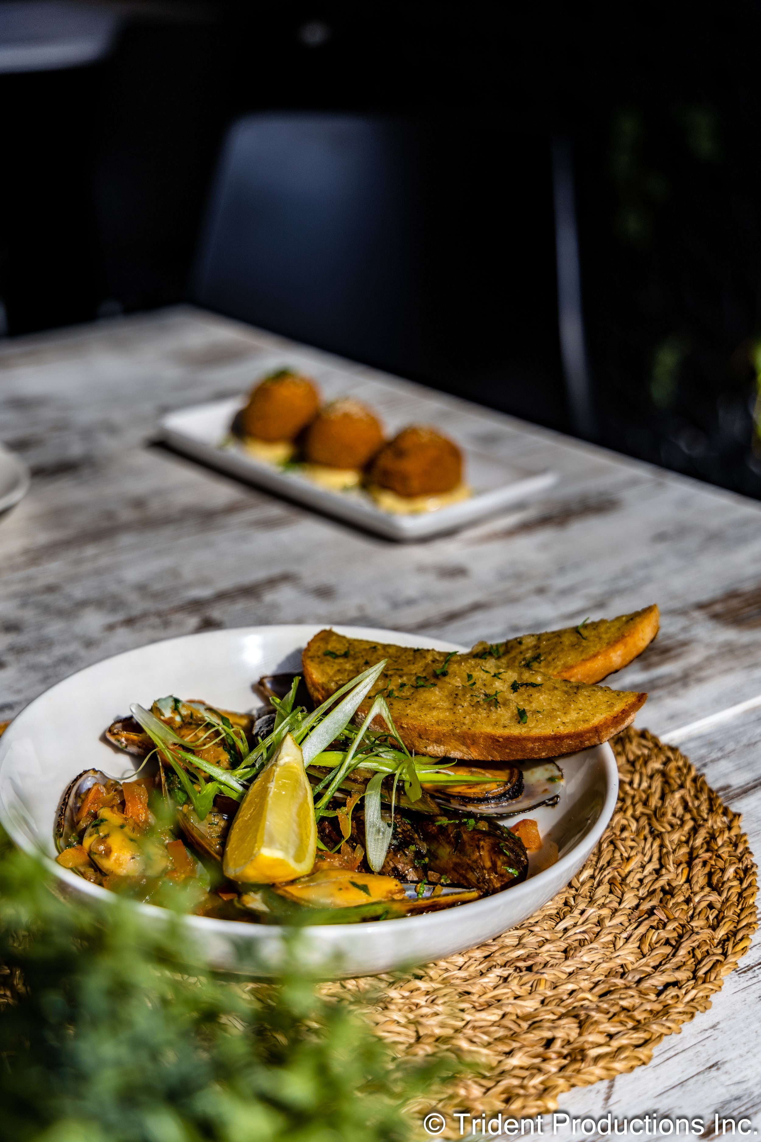 A white bowl of mussels garnished with green herbs, lemon wedge, and a piece of garlic bread on a woven placemat, with three fried appetizers on a rectangular white plate in the background.