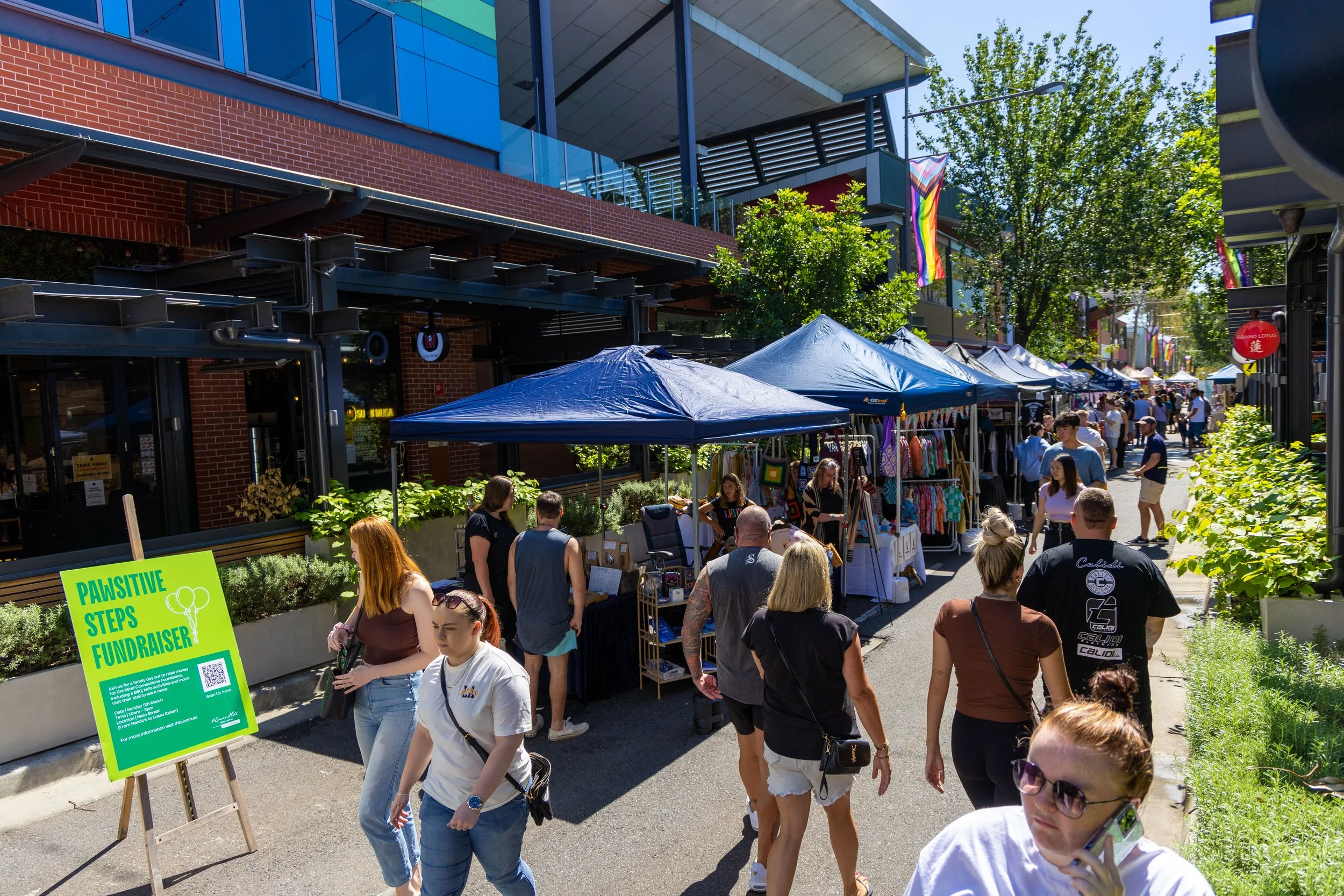 People walking and shopping at outdoor market with vendor tents along a city street, green trees, and colorful flags.