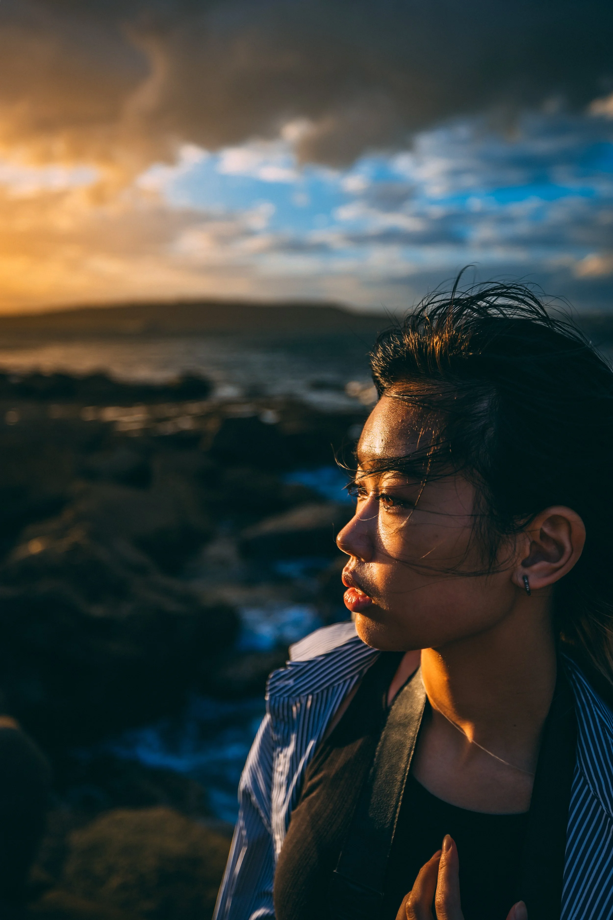 A woman with windblown hair looking towards the sunset by the ocean, with dark clouds and a distant land in the background.