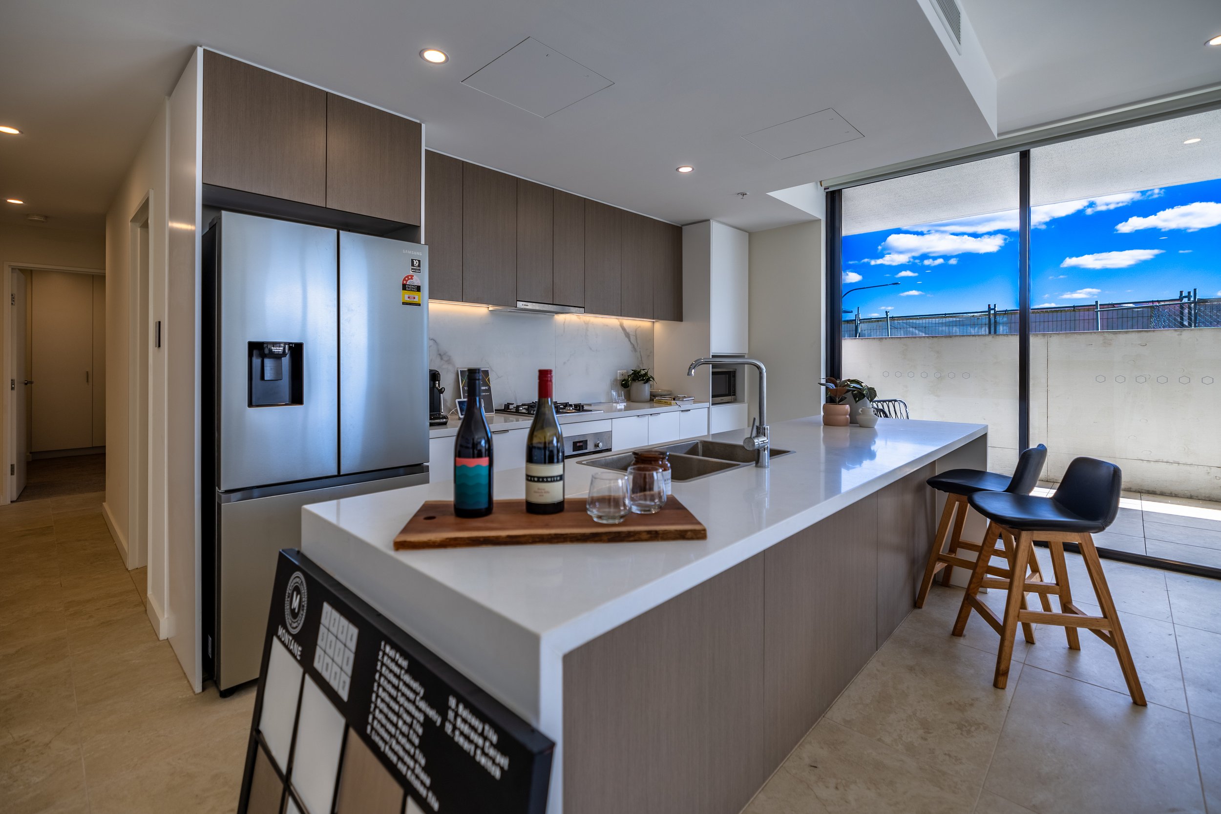 Modern kitchen with white island and bar stools, stainless steel refrigerator, and large sliding glass door showing a blue sky with clouds.