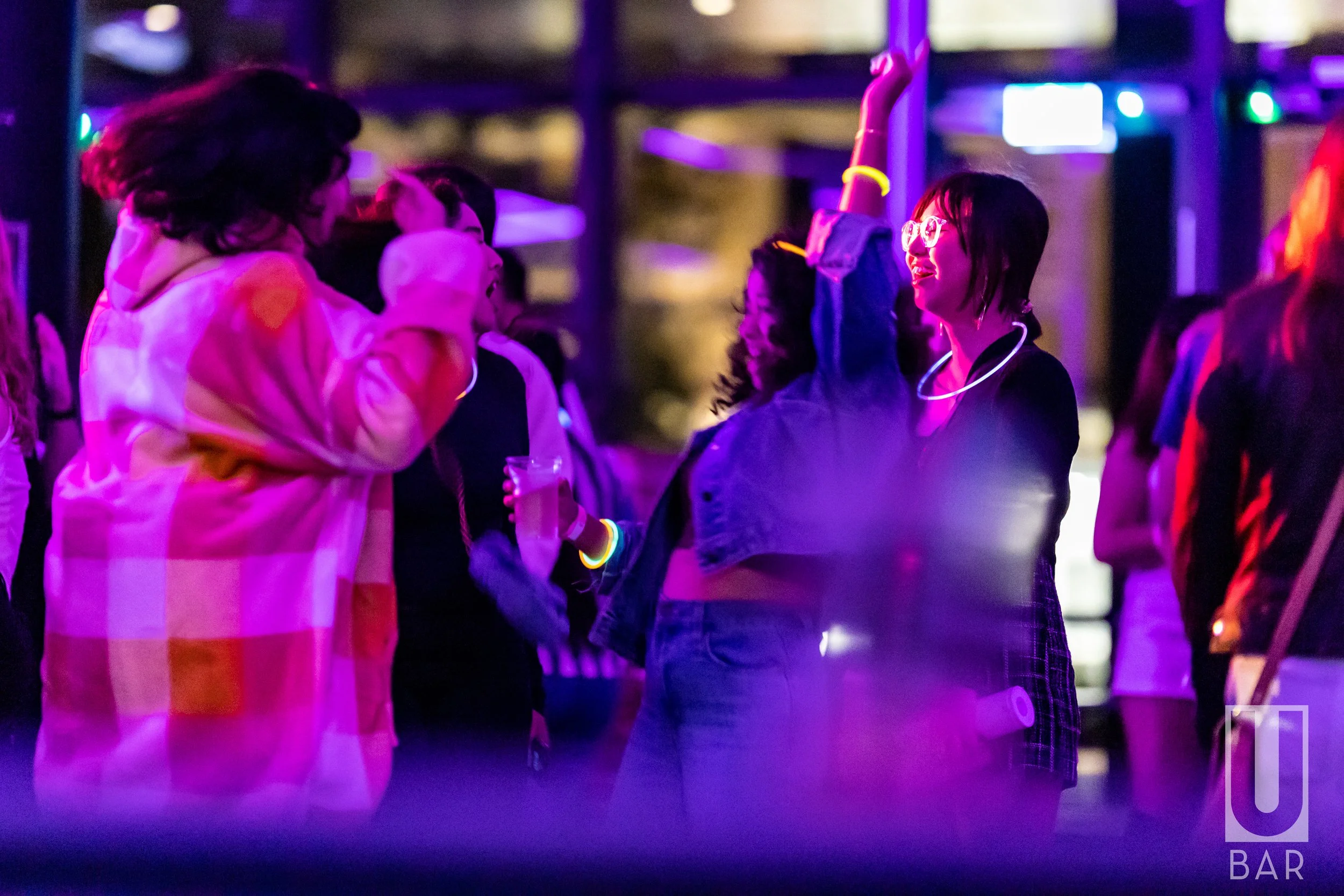 People dancing and socializing at a neon-lit bar or nightclub with purple and pink lighting.