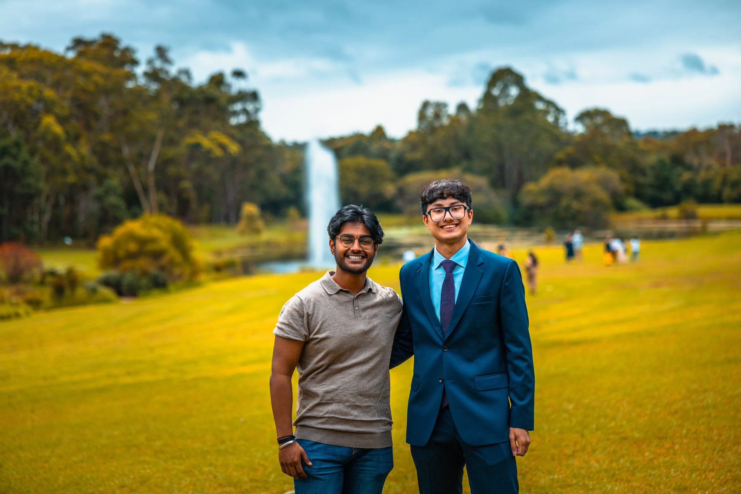 Two men standing together outdoors in front of a waterfall with trees and grass in the background. One man is wearing a casual beige polo shirt and jeans, while the other is dressed in a formal dark blue suit and tie. Both are smiling at the camera.