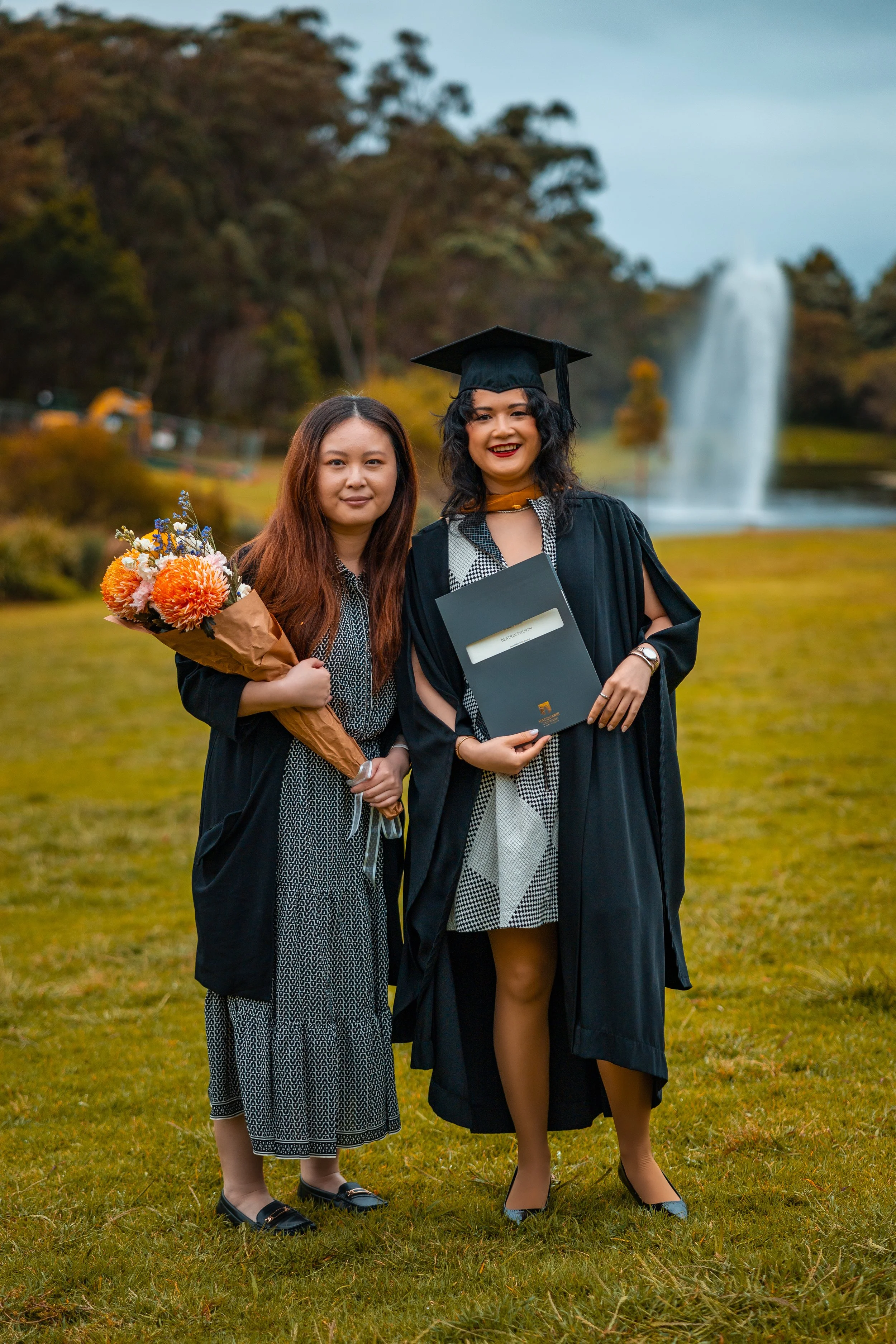 Two women, one in a graduation cap and gown holding a diploma, standing on grass with a waterfall and trees in the background, celebrating graduation.