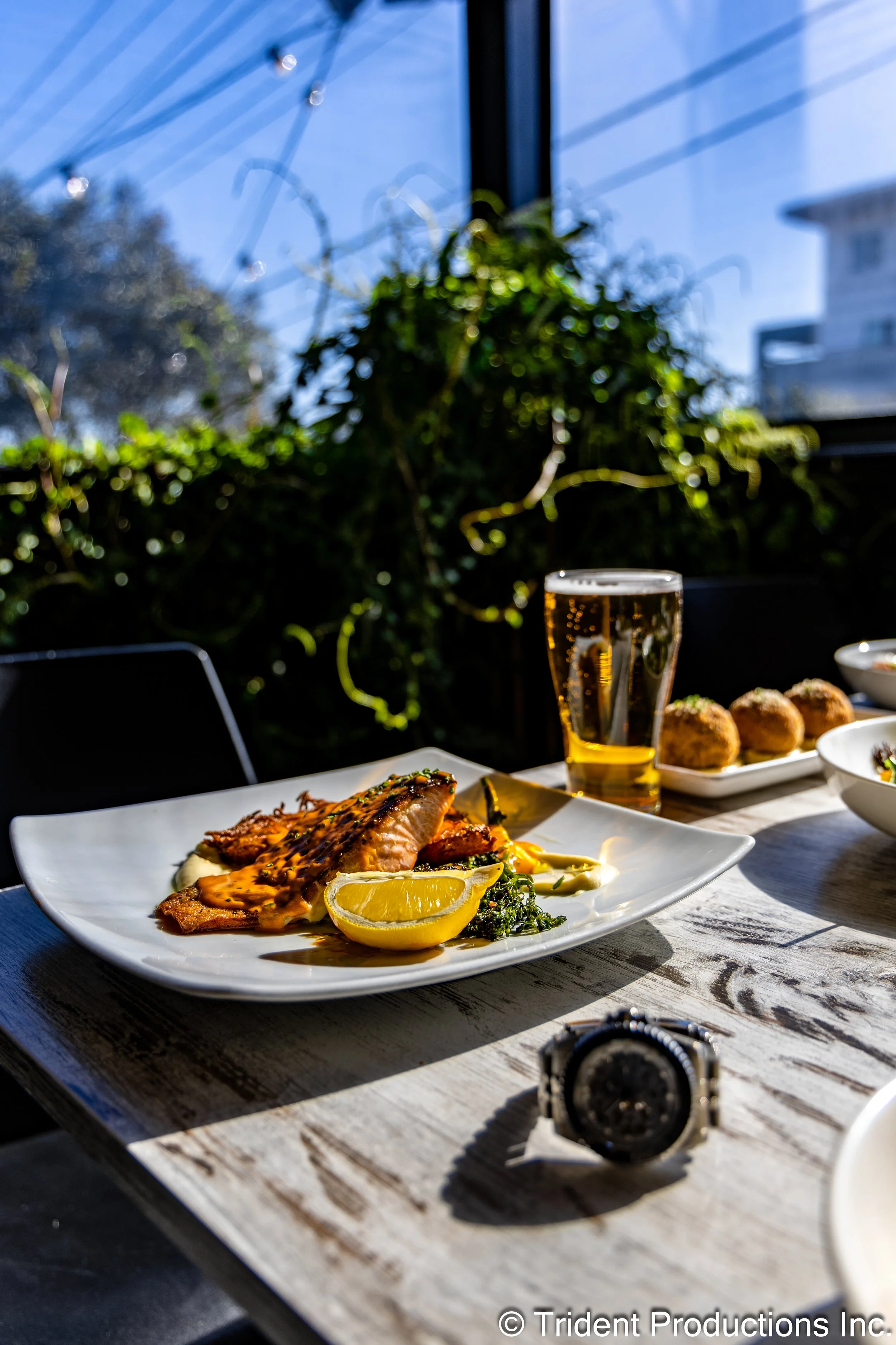 A table with a plate of grilled fish with lemon slices, a glass of beer, and other dishes in the background, set near a window with a sunny outdoor scene.