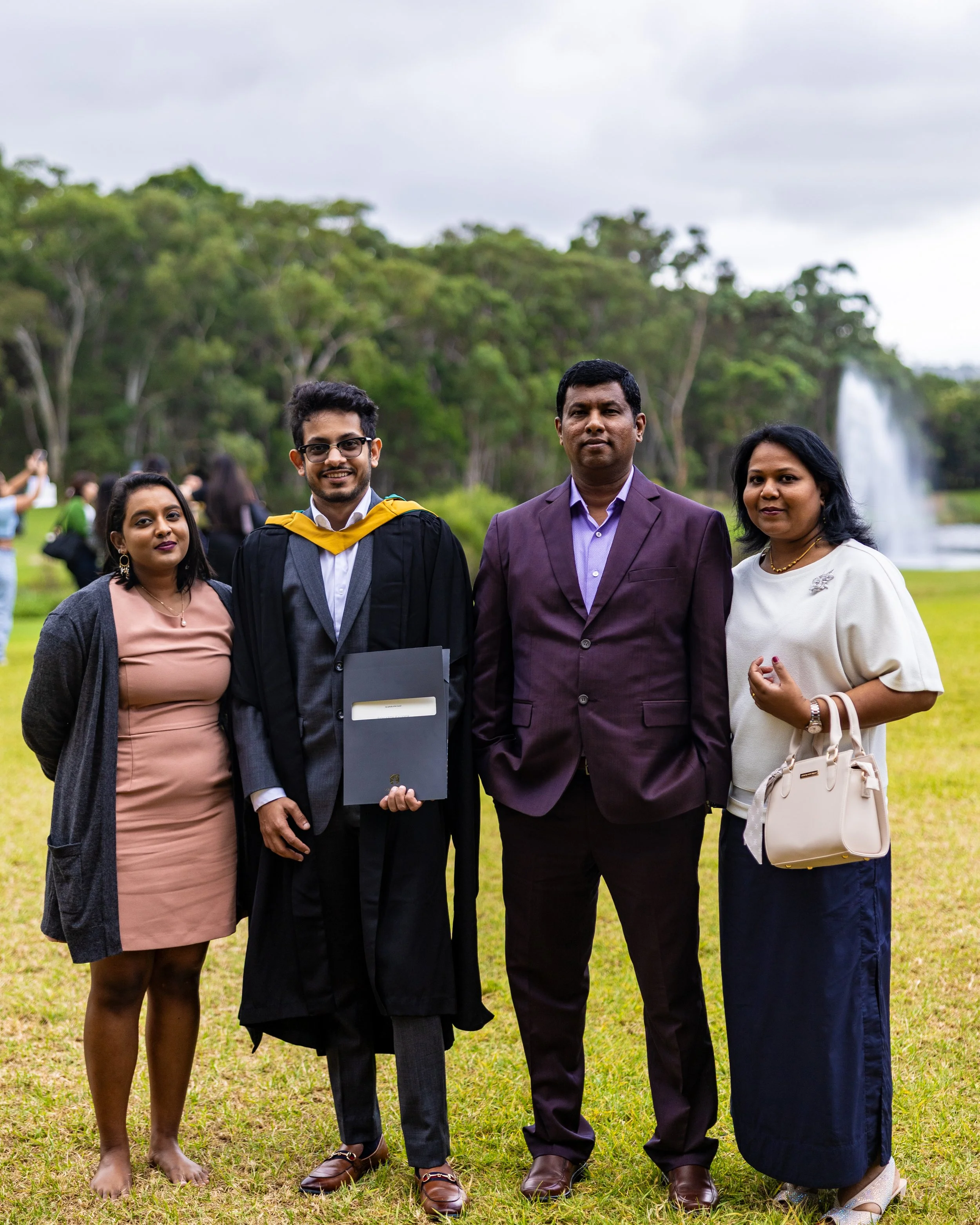 Group of four people standing outdoors during daytime, celebrating a graduation with a fountain and trees in the background.