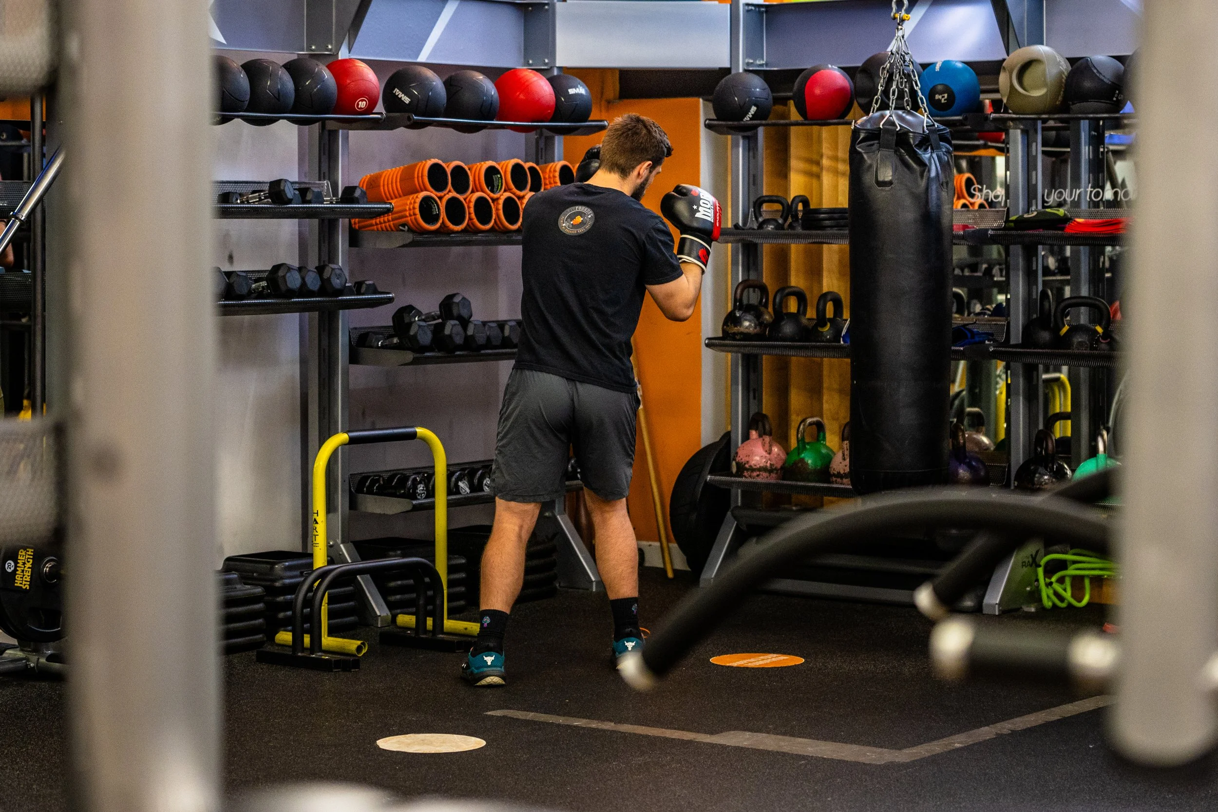 A man in athletic clothing and boxing gloves practicing boxing in a gym with various equipment including kettlebells, medicine balls, dumbbells, and a punching bag.