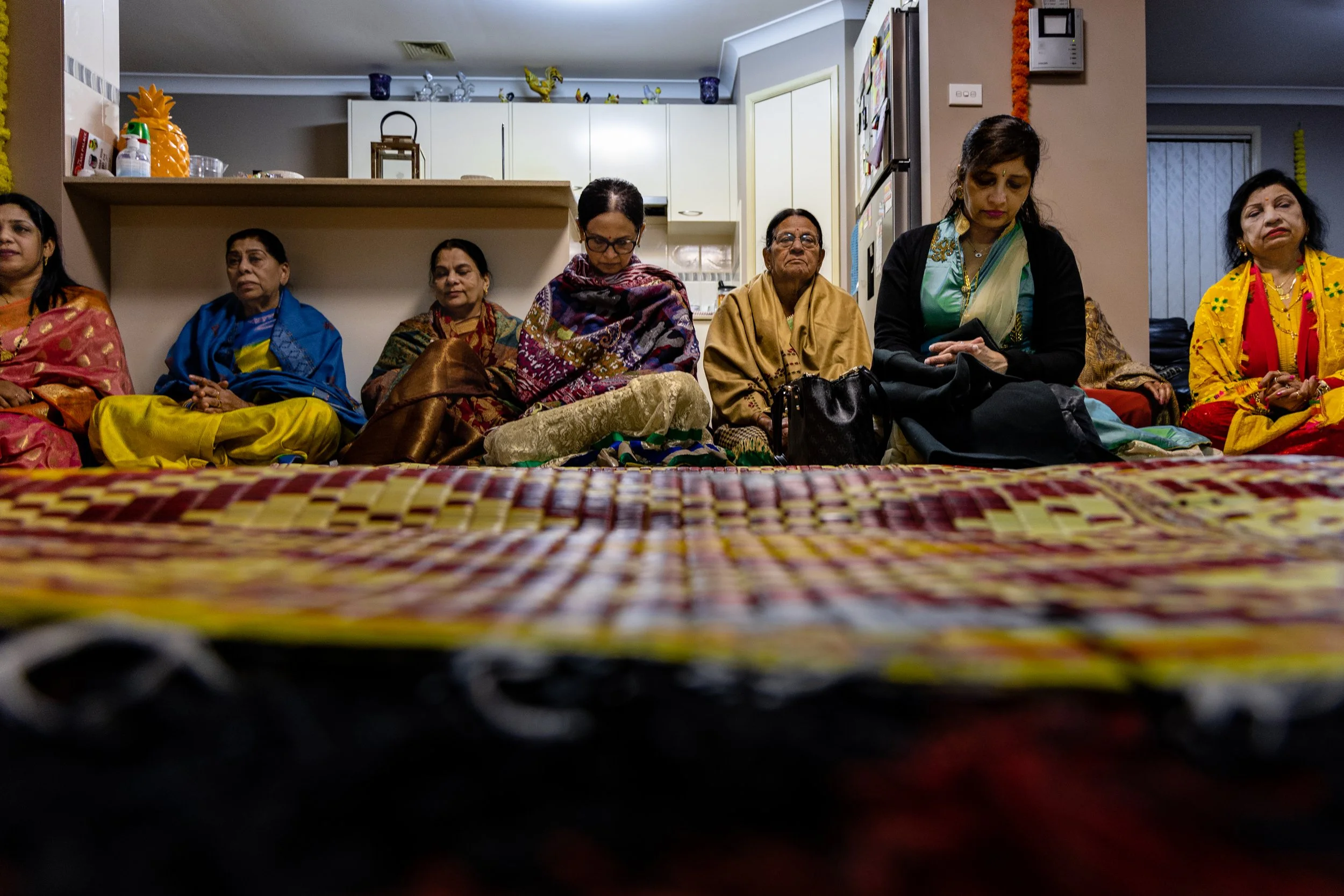 Group of nine women sitting on the floor, dressed in traditional colorful Indian sarees, inside a home, with a kitchen in the background.