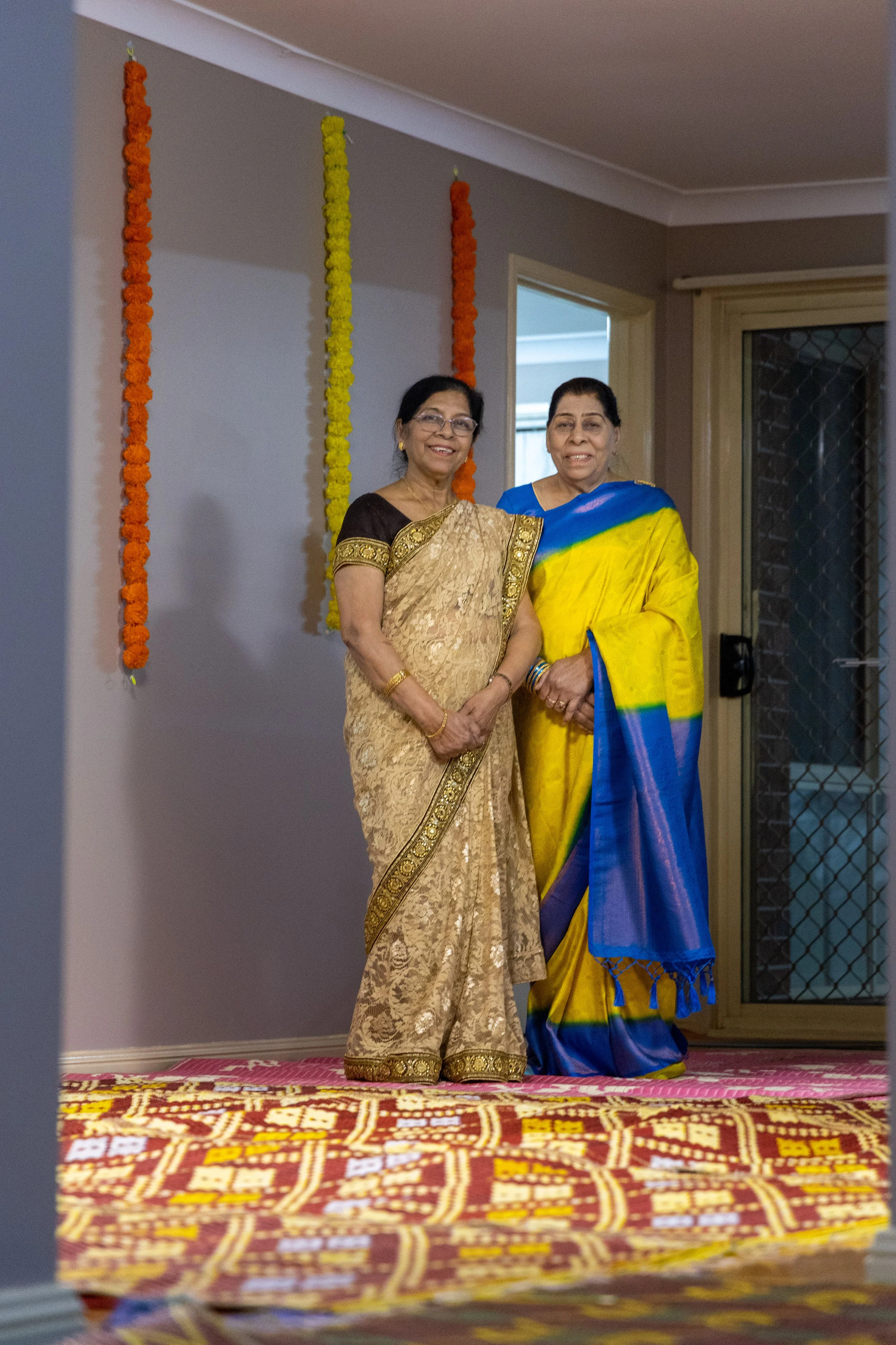 Two women in sarees standing together indoors at a festive or ceremonial event, with colorful marigold flower garlands hanging on the wall behind them.