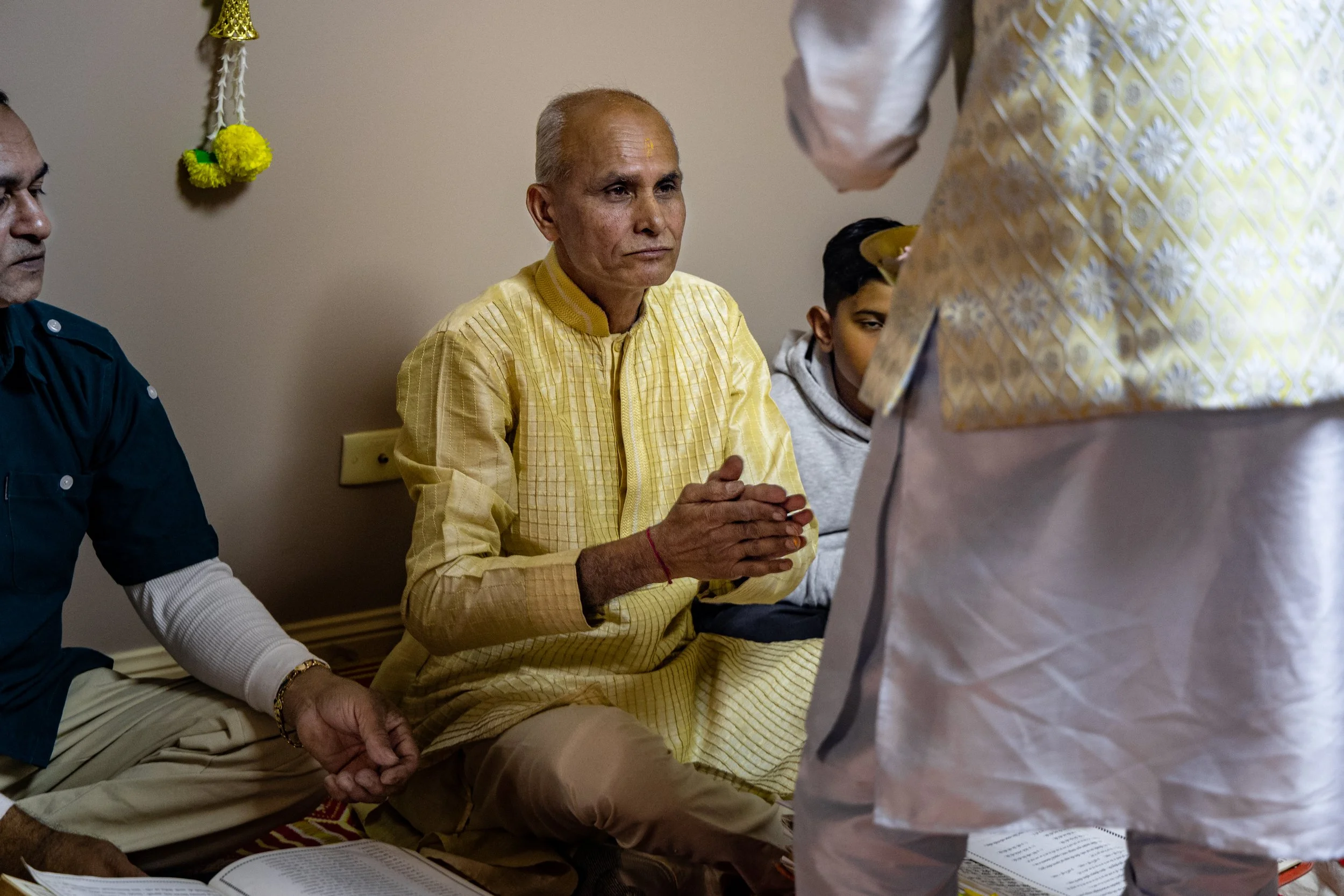 A man in a yellow traditional Indian outfit sitting on the floor with hands folded in prayer, participating in a religious ceremony, with two other men and a boy nearby.