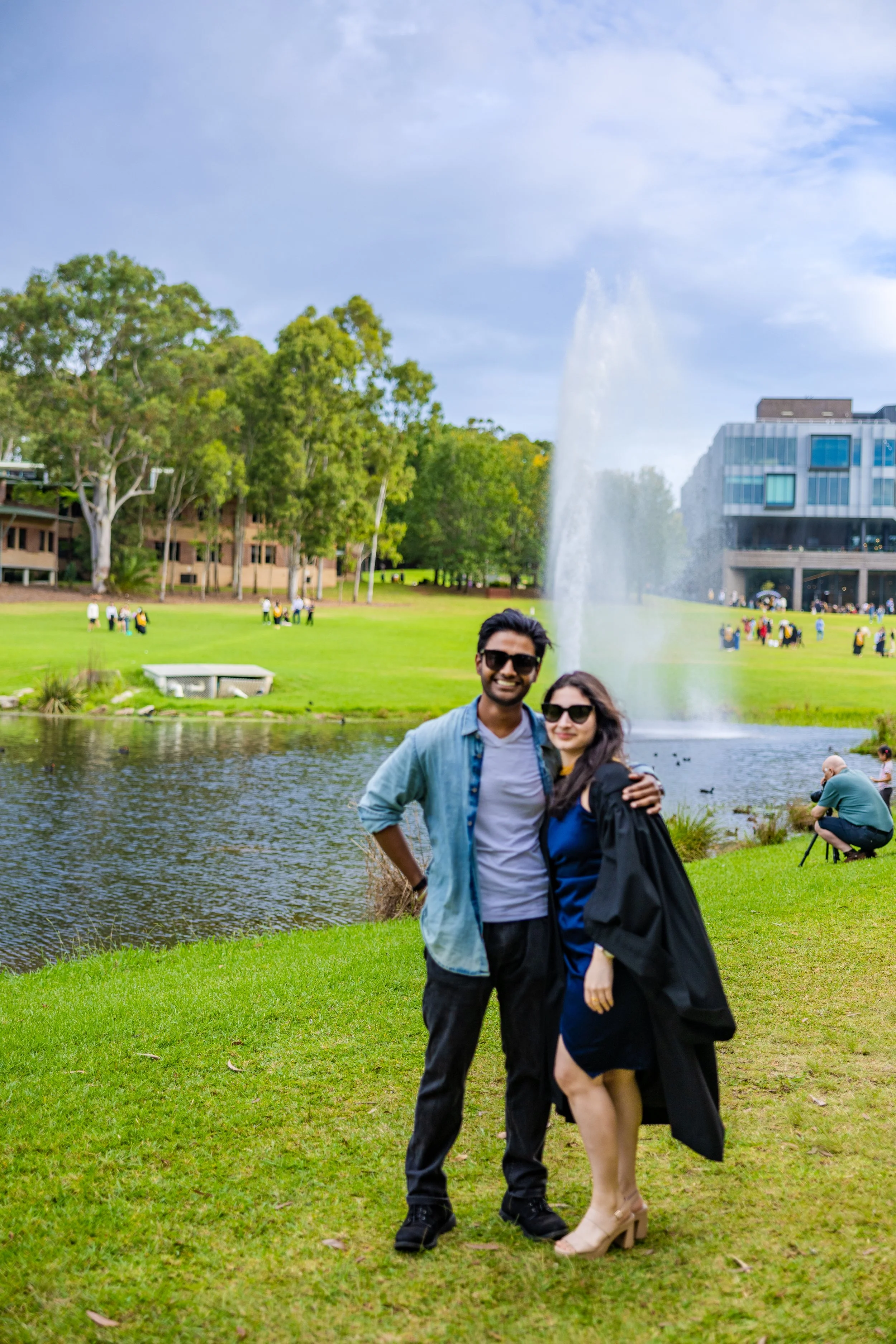 A smiling couple standing near a pond with a fountain in a park, trees, and a modern building in the background.