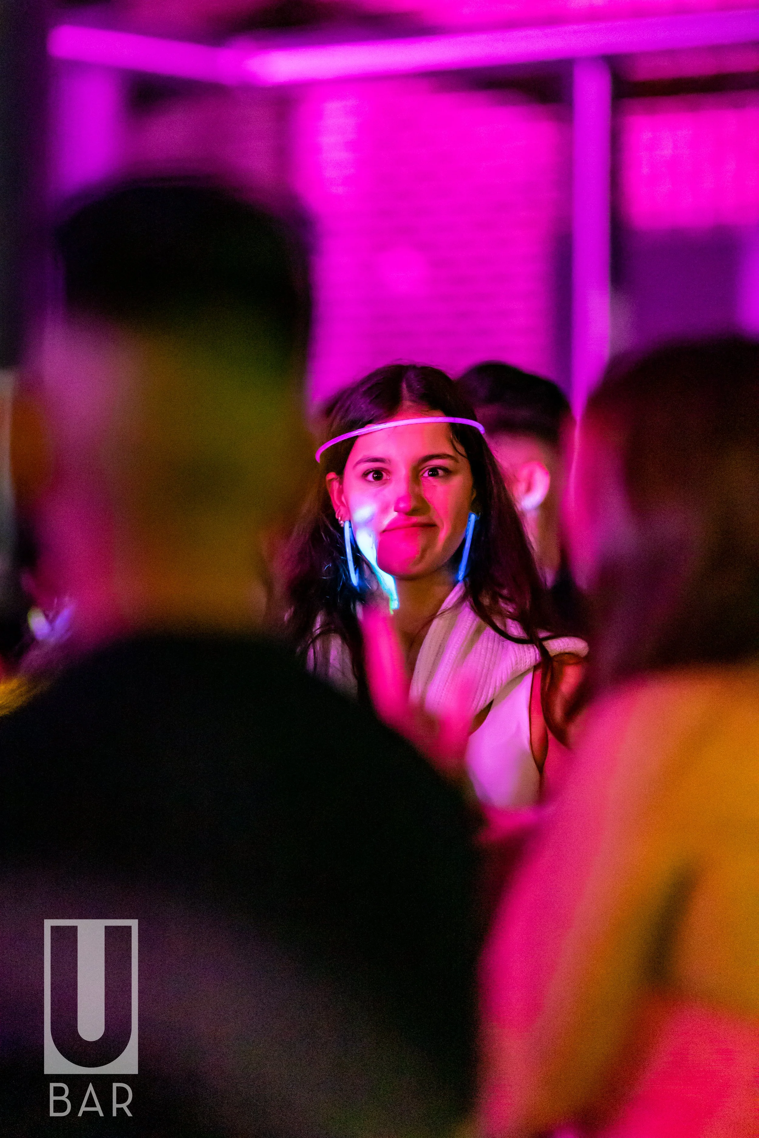 A woman with long dark hair wearing glow-in-the-dark earrings and a glow headband, standing in a bar with pink and purple lighting, surrounded by a crowd of people.