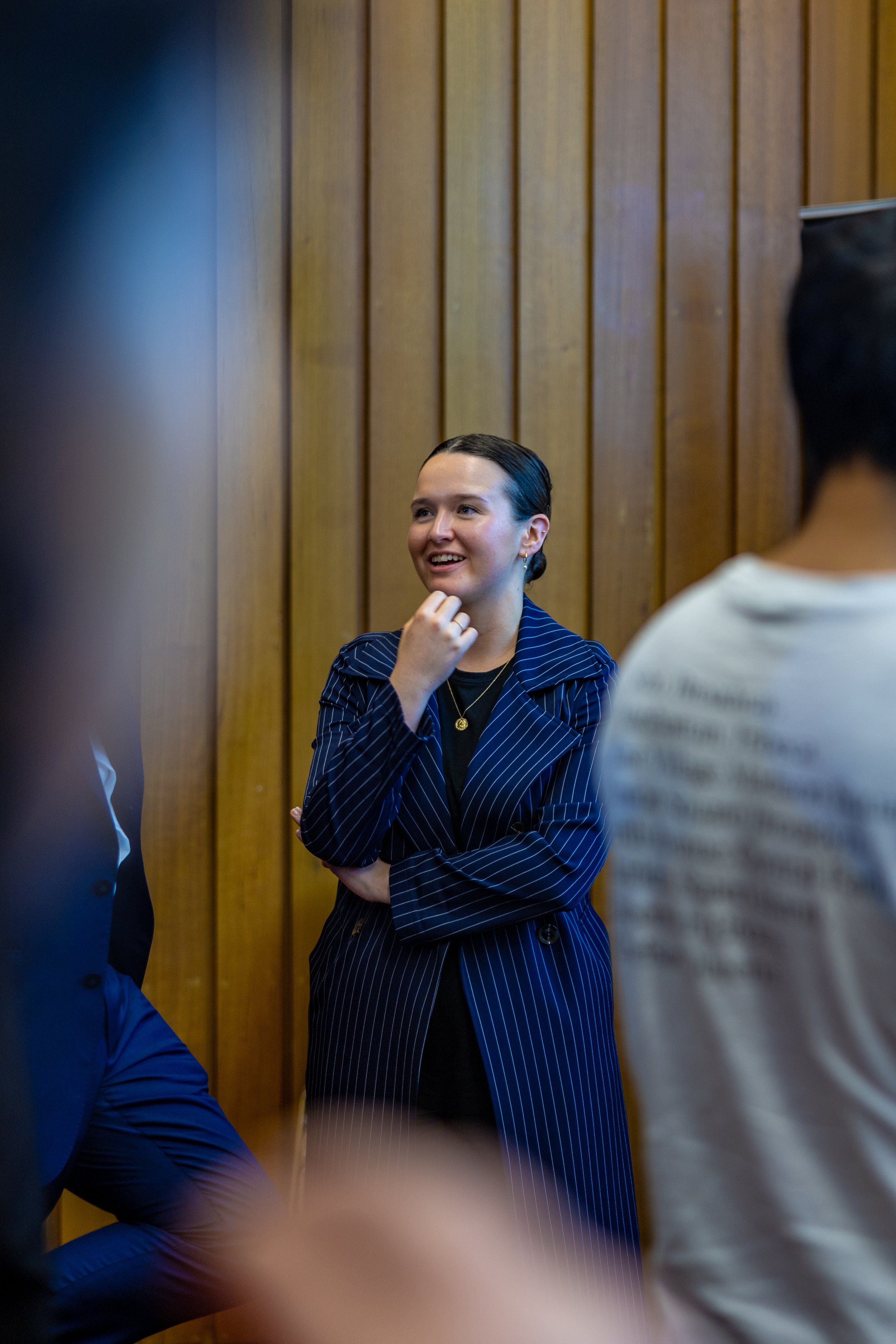 A woman with dark, slicked-back hair, wearing a navy blue pinstripe blazer, stands with her hand on her chin, smiling during a conversation in a room with wooden wall paneling, surrounded by other people.