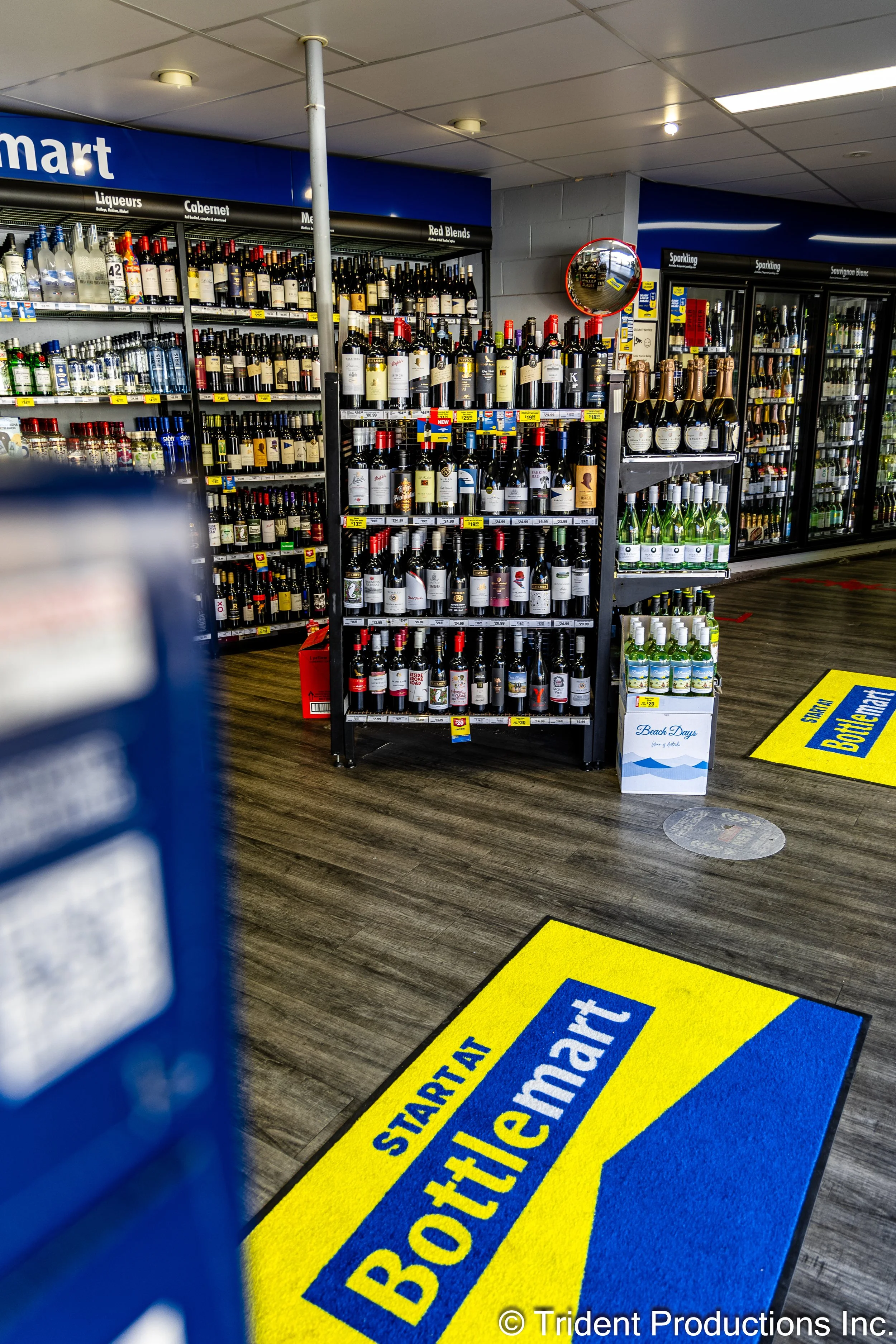 A store aisle with a Bottlemart start mat on the wooden floor, shelf displays of various wines and spirits, and a mirror on the wall reflecting the store interior.