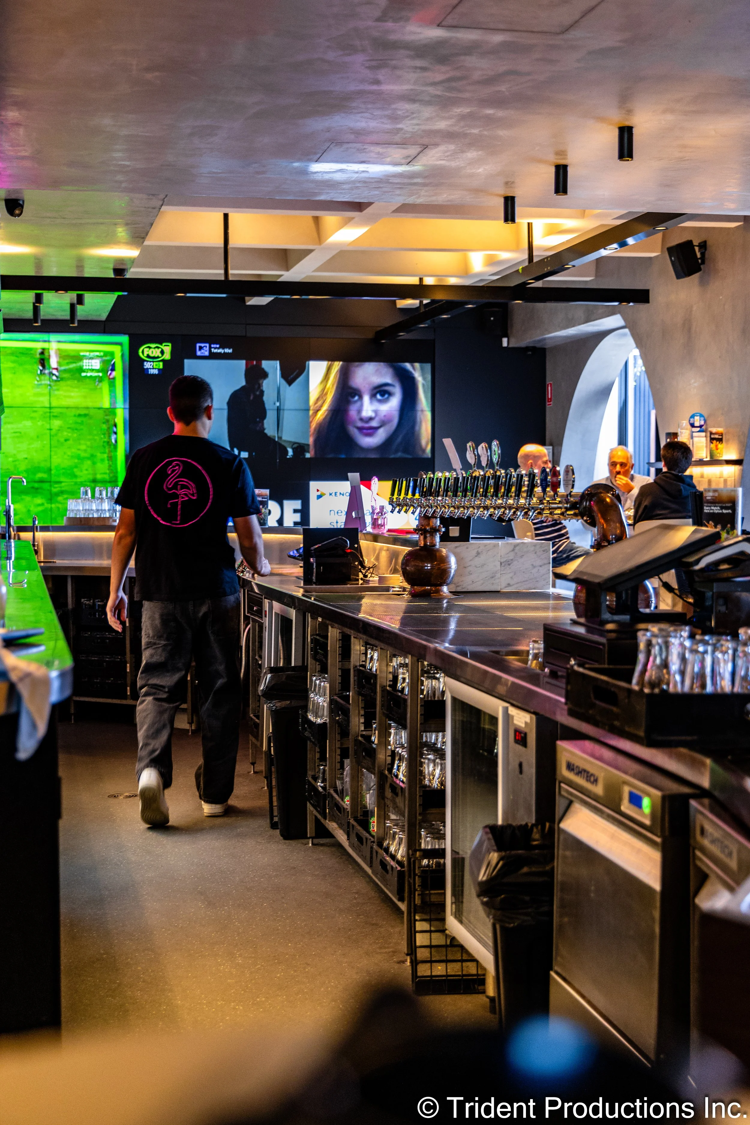 Interior of a sports bar with a bartender walking past a counter with beer taps, surrounded by screens displaying sports and a woman's face.