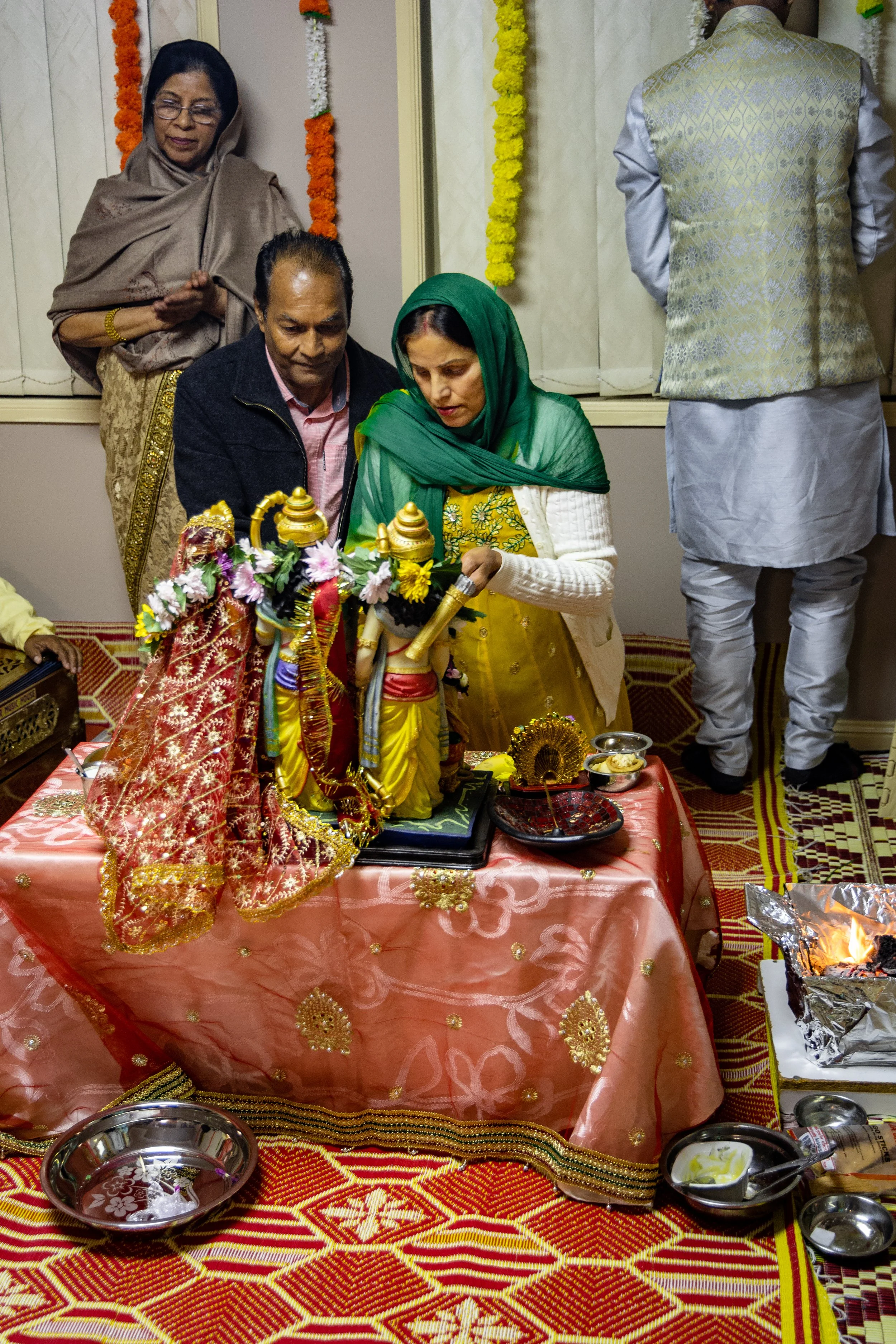 People participating in a Hindu religious ceremony, worshipping deities on a decorated table, with offerings and ritual items.