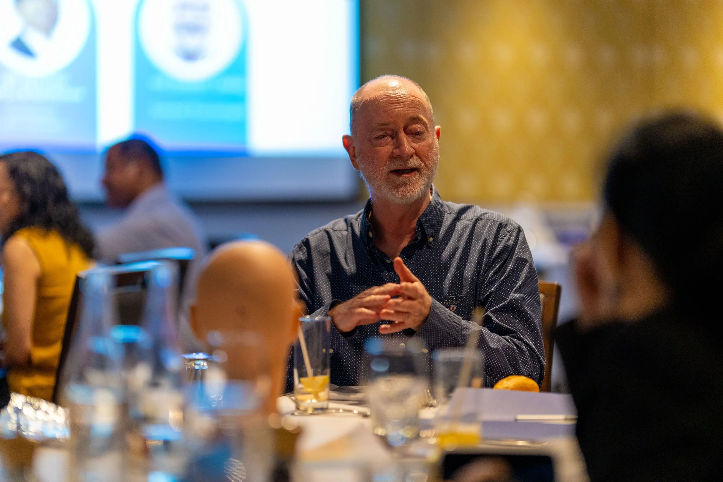 Man with gray hair and beard speaking during a meeting at a round table in a conference room, with others sitting around.