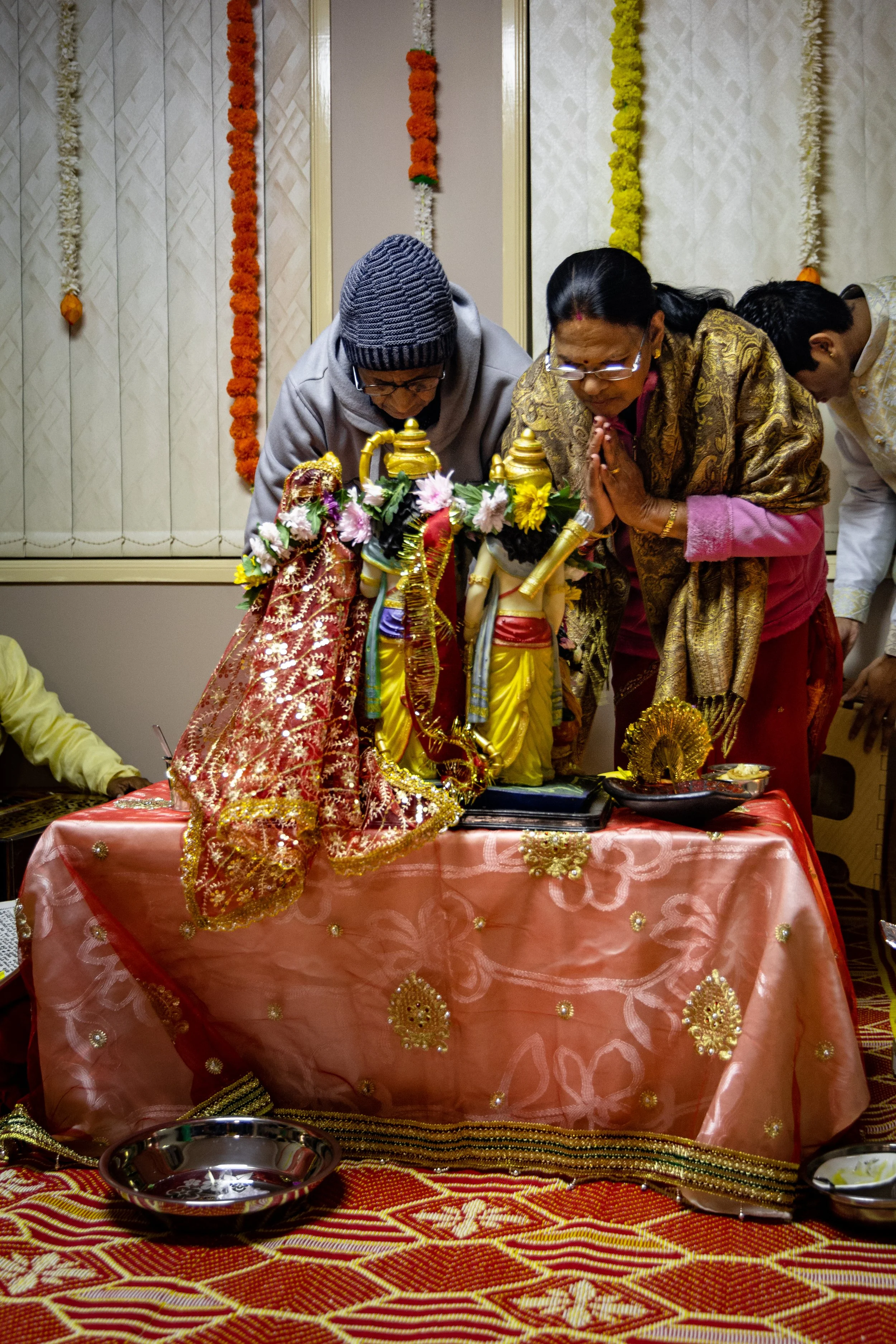 People performing a religious ritual in front of decorated idols and statues on a table draped with a pink cloth, adorned with flowers and ornaments, in a festive setting.