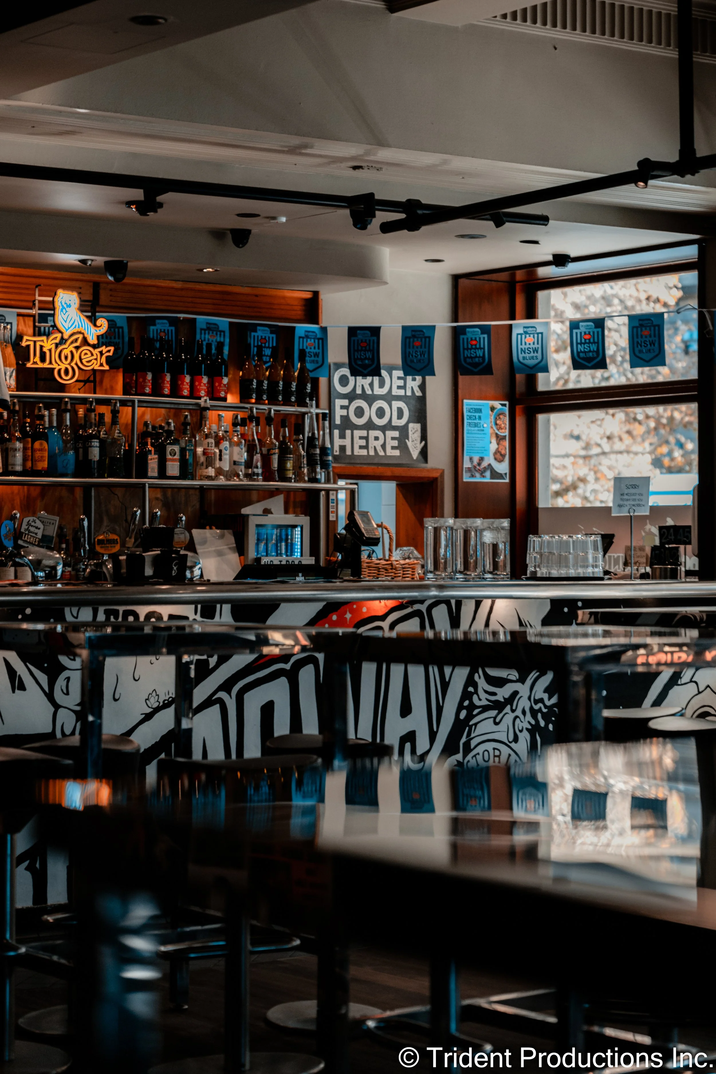 Empty bar and seating area inside a restaurant or bar, with a sign that says 'Order Food Here' and a decorated bar counter with bottles of alcohol, glasses, and a neon sign that says 'Tiger.' There are windows with outdoor light and blue banners hang