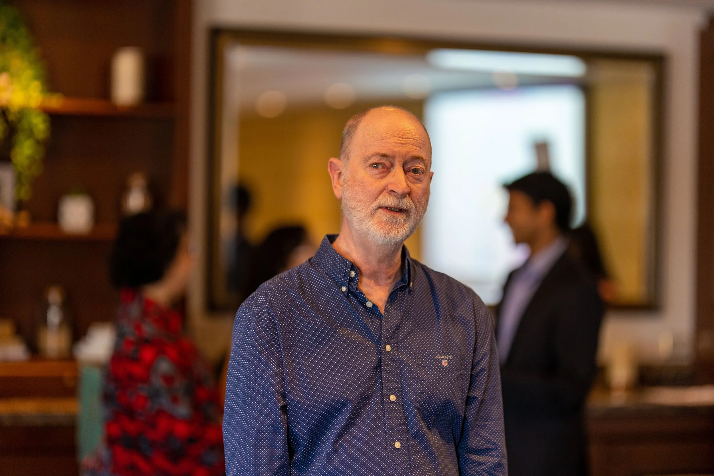 An older man with a beard and mustache wearing a blue button-up shirt standing in a warmly lit room with blurred people in the background.