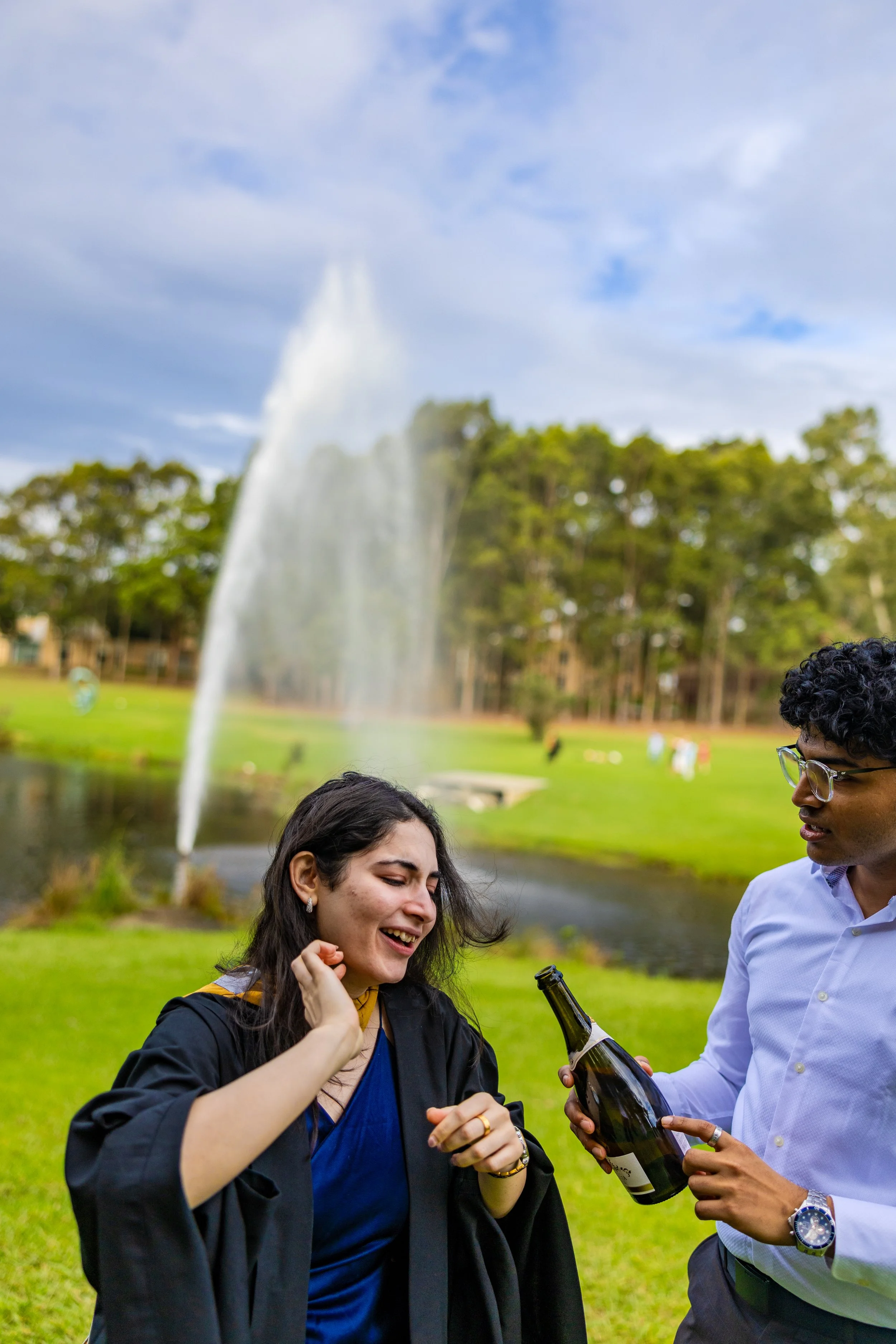 A young woman in a graduation gown smiling and laughing as a young man offers her a bottle of champagne in a park with a fountain and trees in the background.