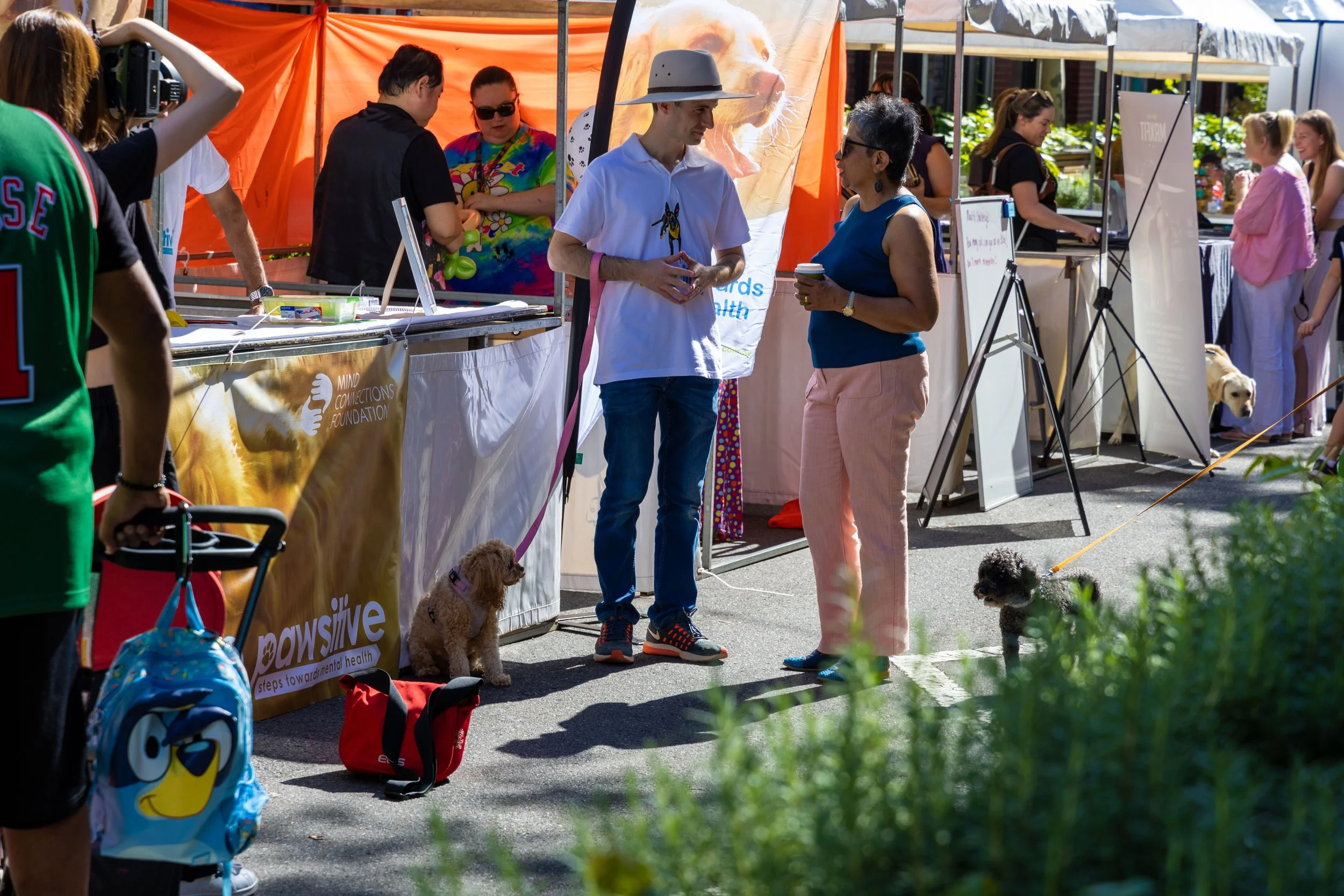 People walking and talking at an outdoor event with booths and tents. Two women are in conversation, one holding a small dog on a leash. Several dogs and children are present. The scene is sunny with bright colors and various attendees.