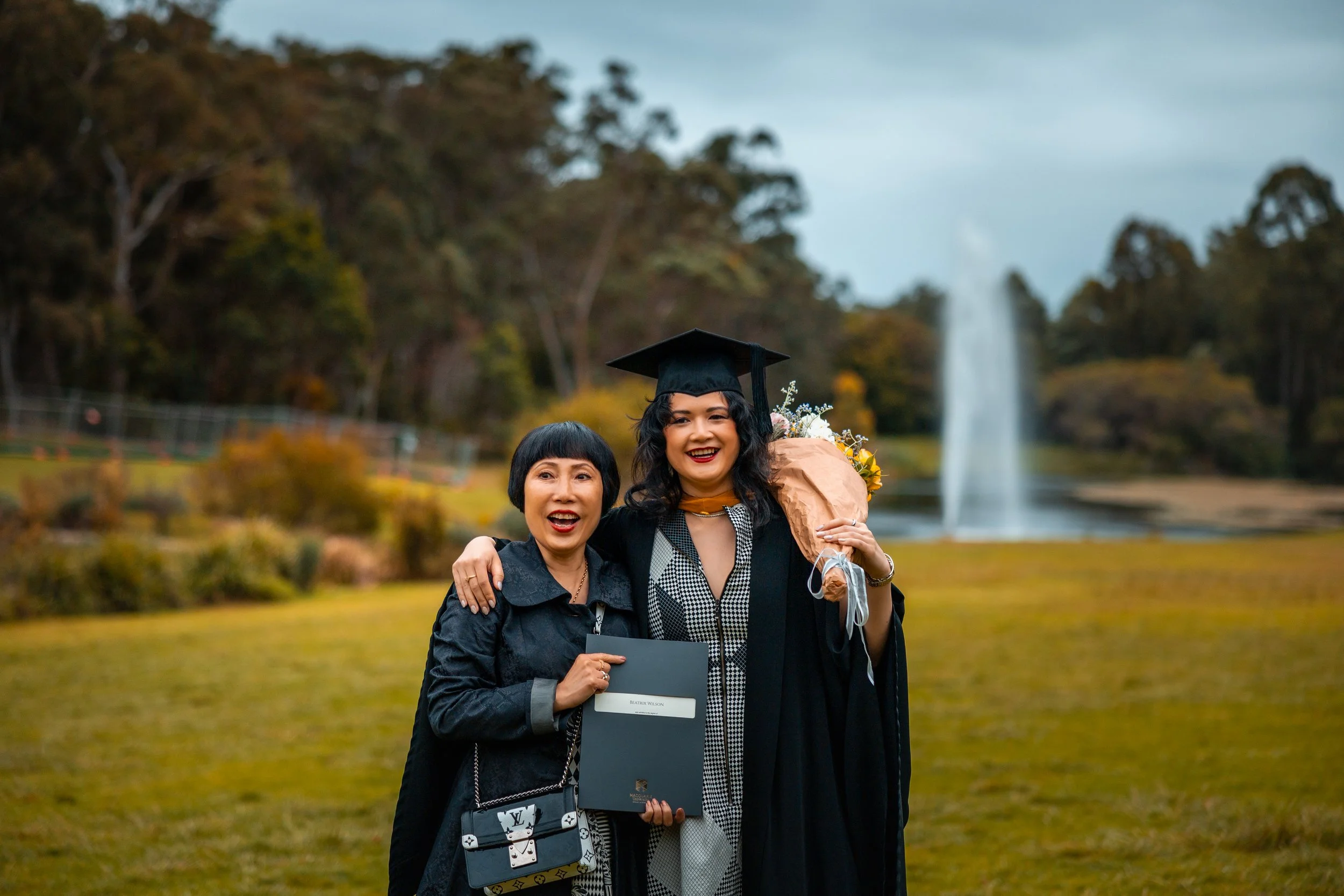 A woman in a graduation gown and cap holding a bouquet of flowers standing next to another woman in black clothing, outdoors near a fountain and trees in the background.