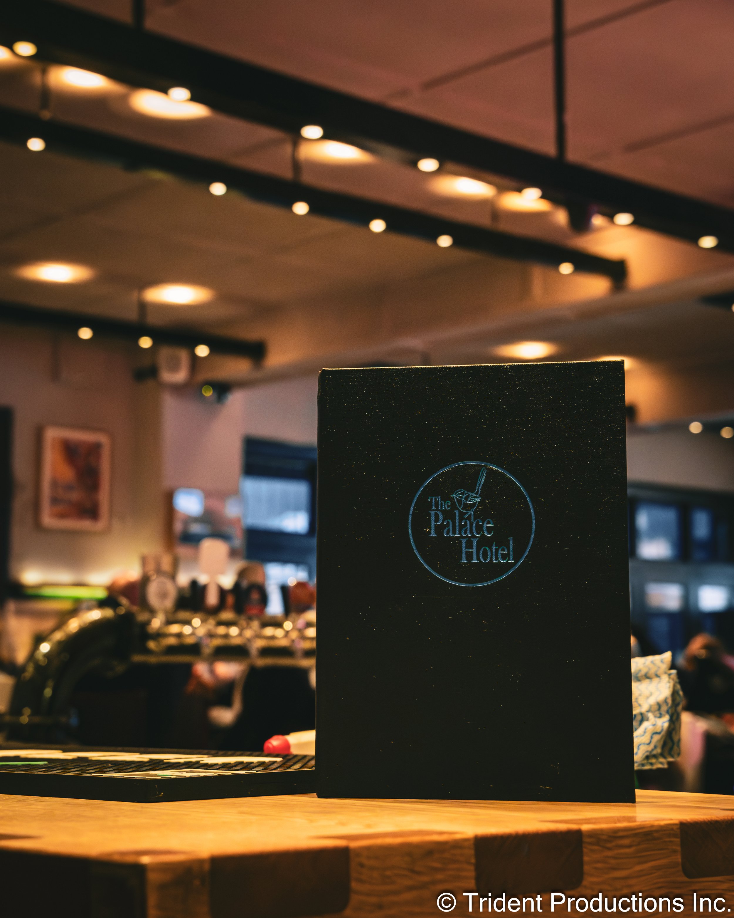 A menu or sign for The Palace Hotel on a table in a dimly lit restaurant or bar with a bar counter and patrons in the background.
