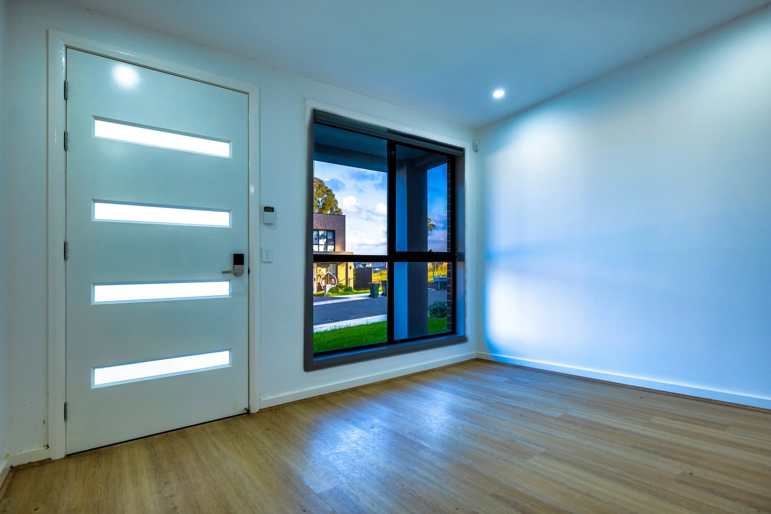 Empty room with wooden floor, white walls, a white front door with horizontal window slits, and a large window showing a street view.