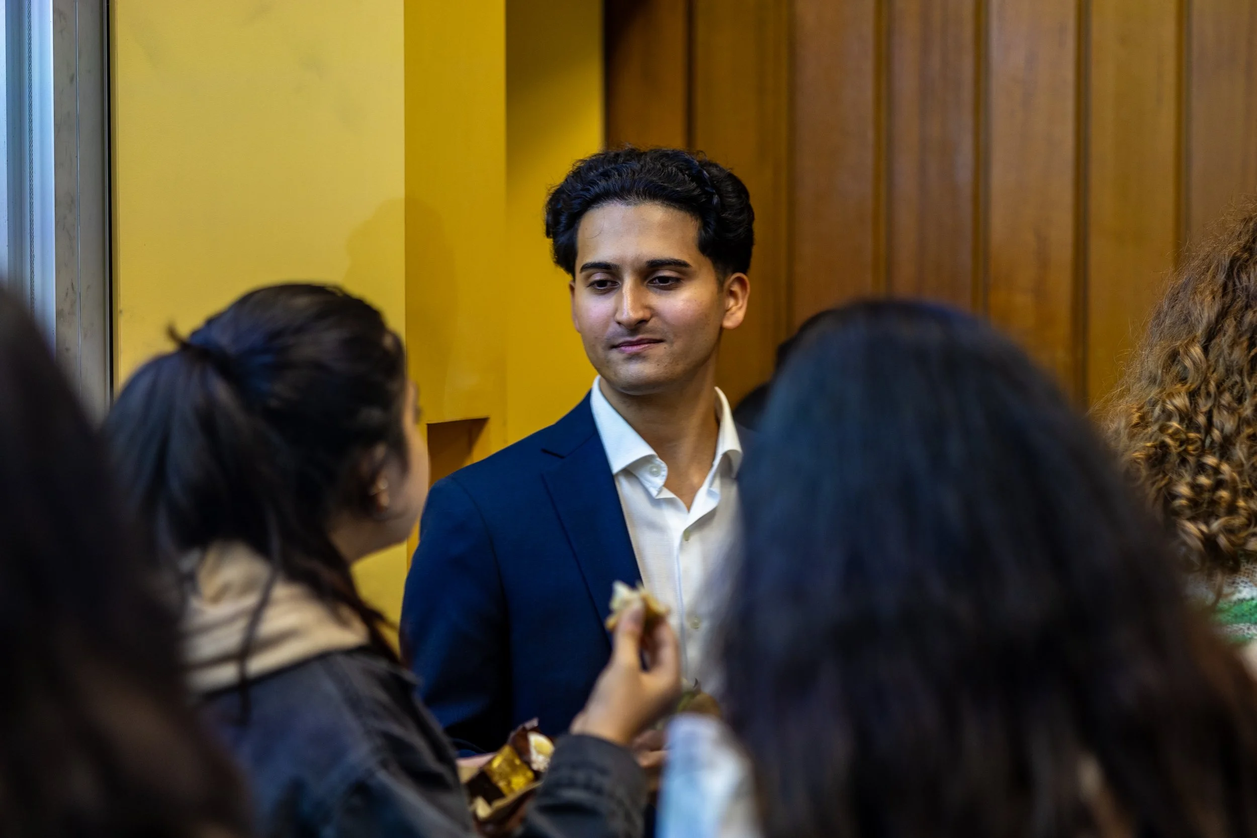 Man in a navy blue suit and white shirt talking to a group of women in a room with yellow and wooden walls.