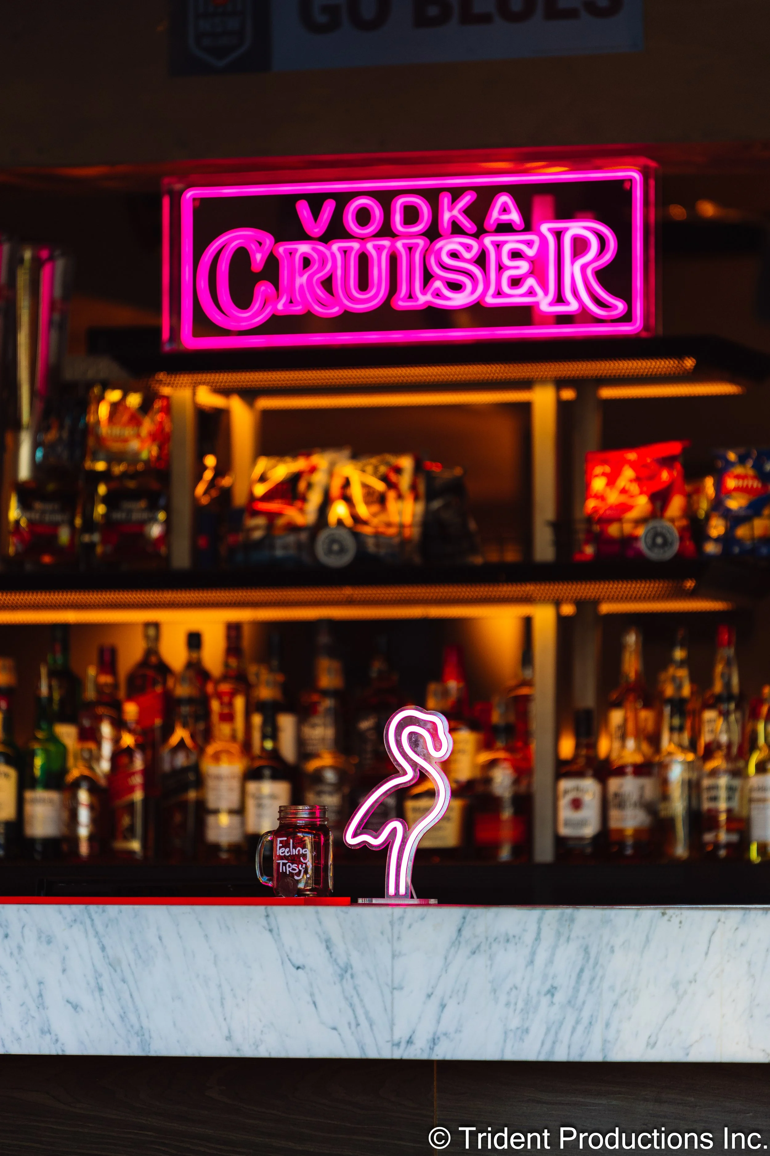 Bright pink neon sign that reads 'VODKA CRUISER' hanging above a bar counter with a flamingo-shaped neon light and a mason jar with the words 'Feeling Tipsy' on it. Shelves with various liquor bottles are in the background.